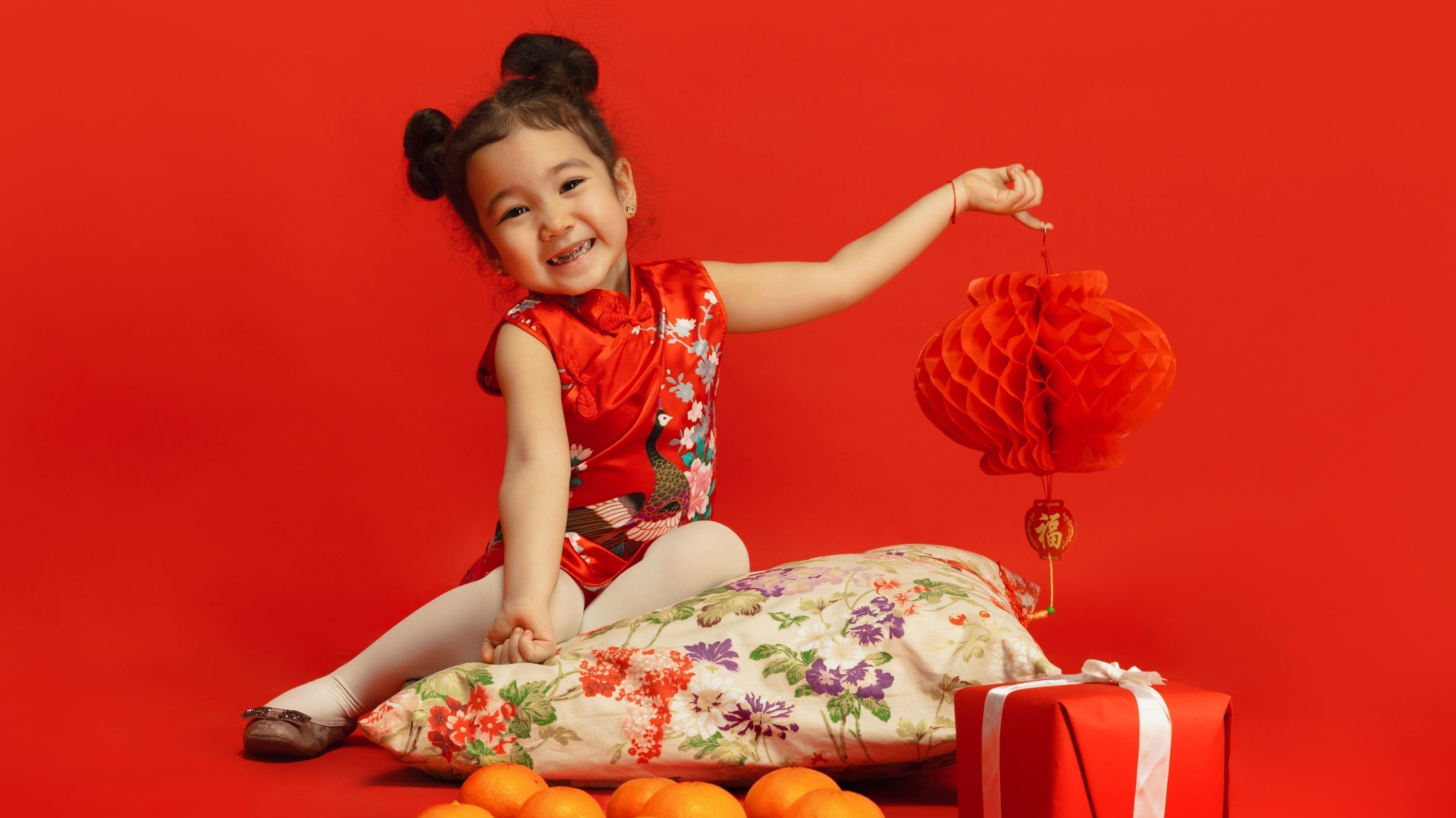 Child celebrating Lunar New Year in red traditional dress holding a lantern with oranges and festive decorations.