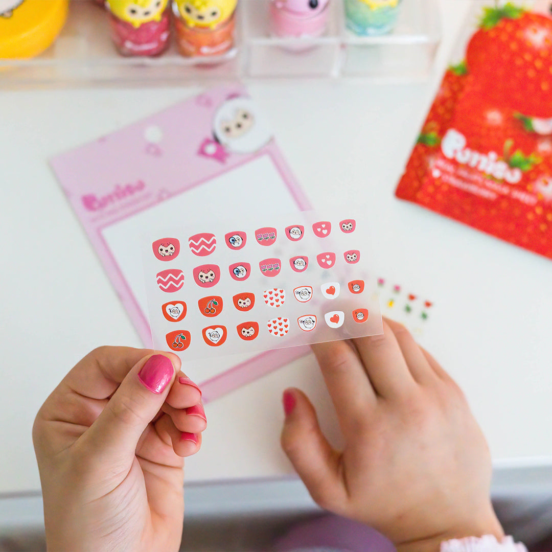 Close-up of a person holding a sheet of nail art stickers with cute designs like owls and hearts, with a pink notepad and other cosmetic items in the background.