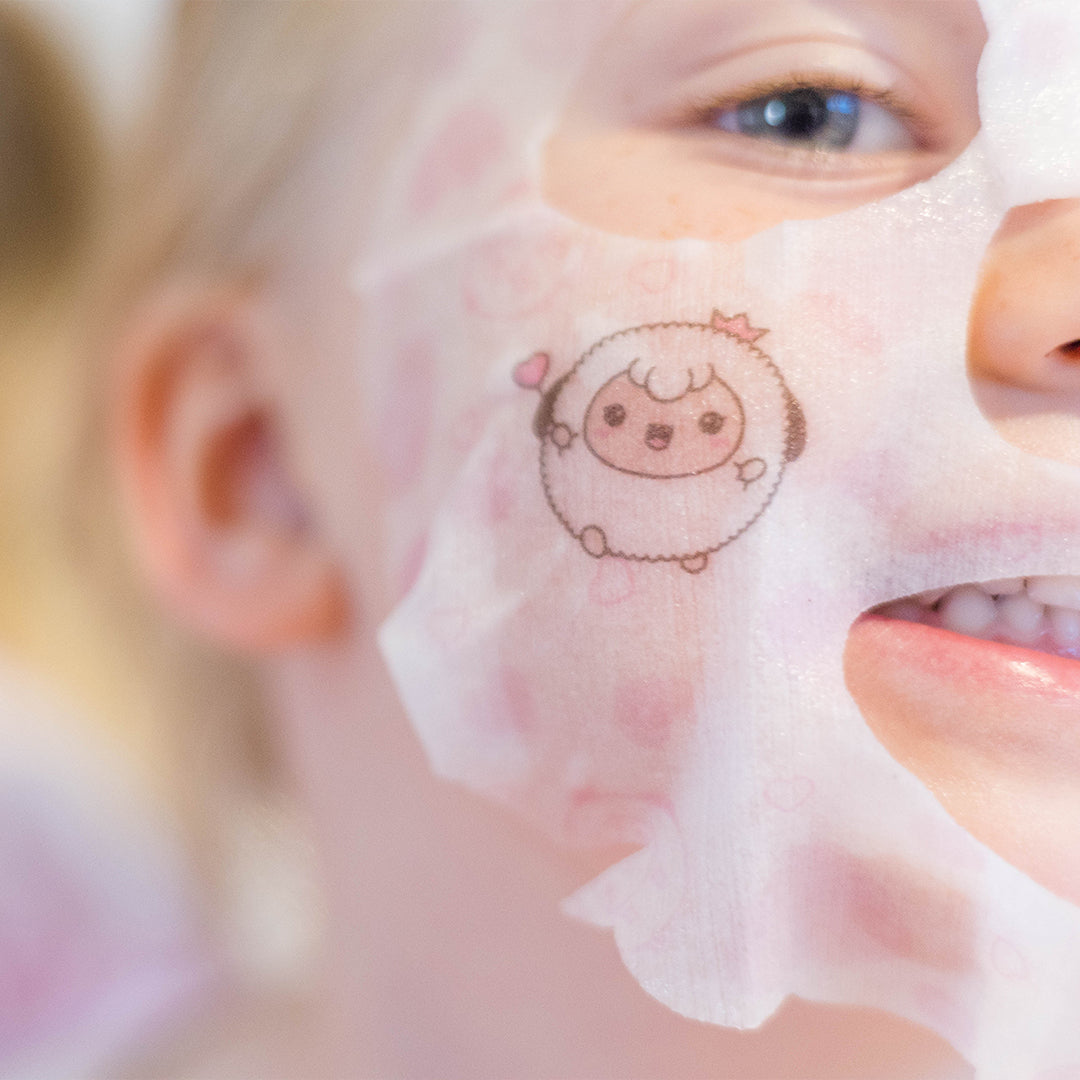 Close-up of a fair-skinned person wearing a sheet face mask with a cartoon sheep design and small pink hearts, showing their blue eye, ear, and teeth.