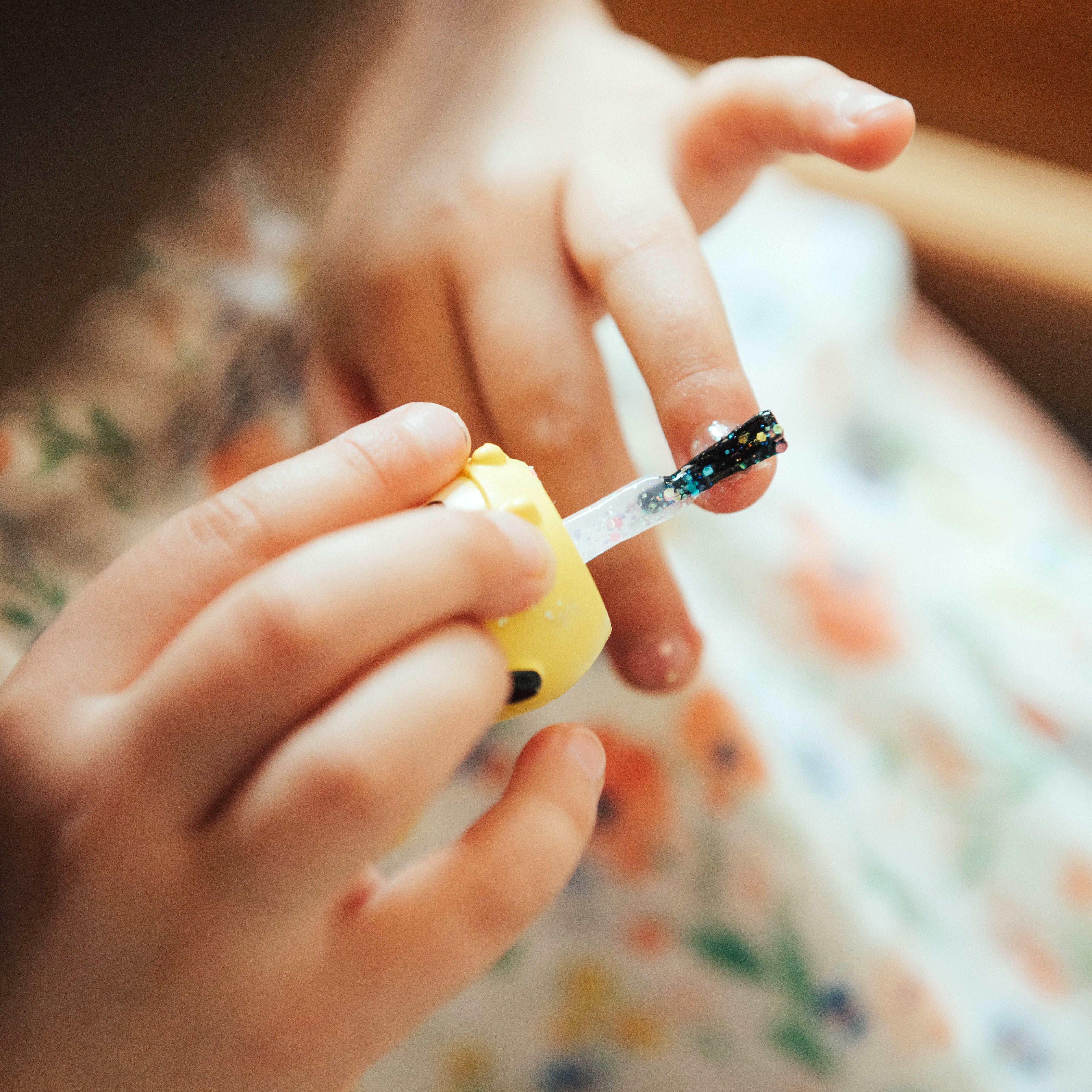 Child applying glitter nail polish on fingernail. Floral dress.