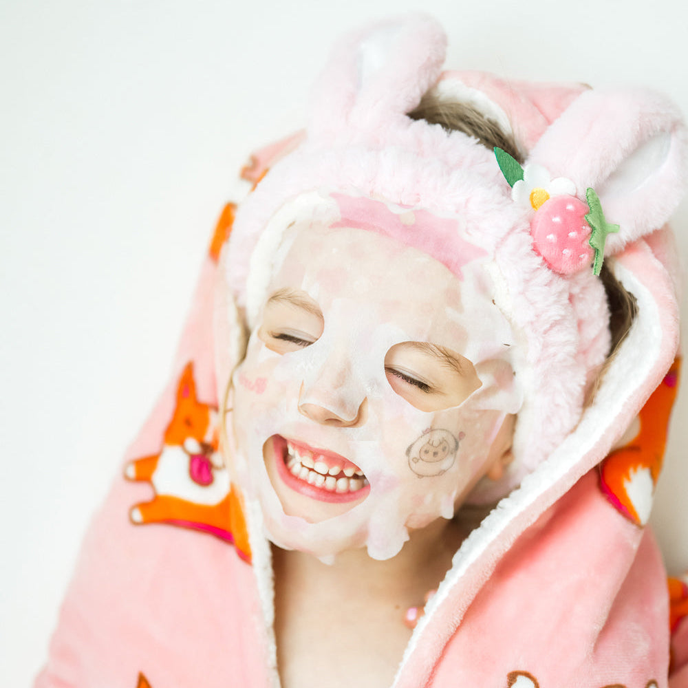 kid wearing a pink plush robe and bunny ear plush spa headband with a Puttisu face sheet mask, smiling against a white background