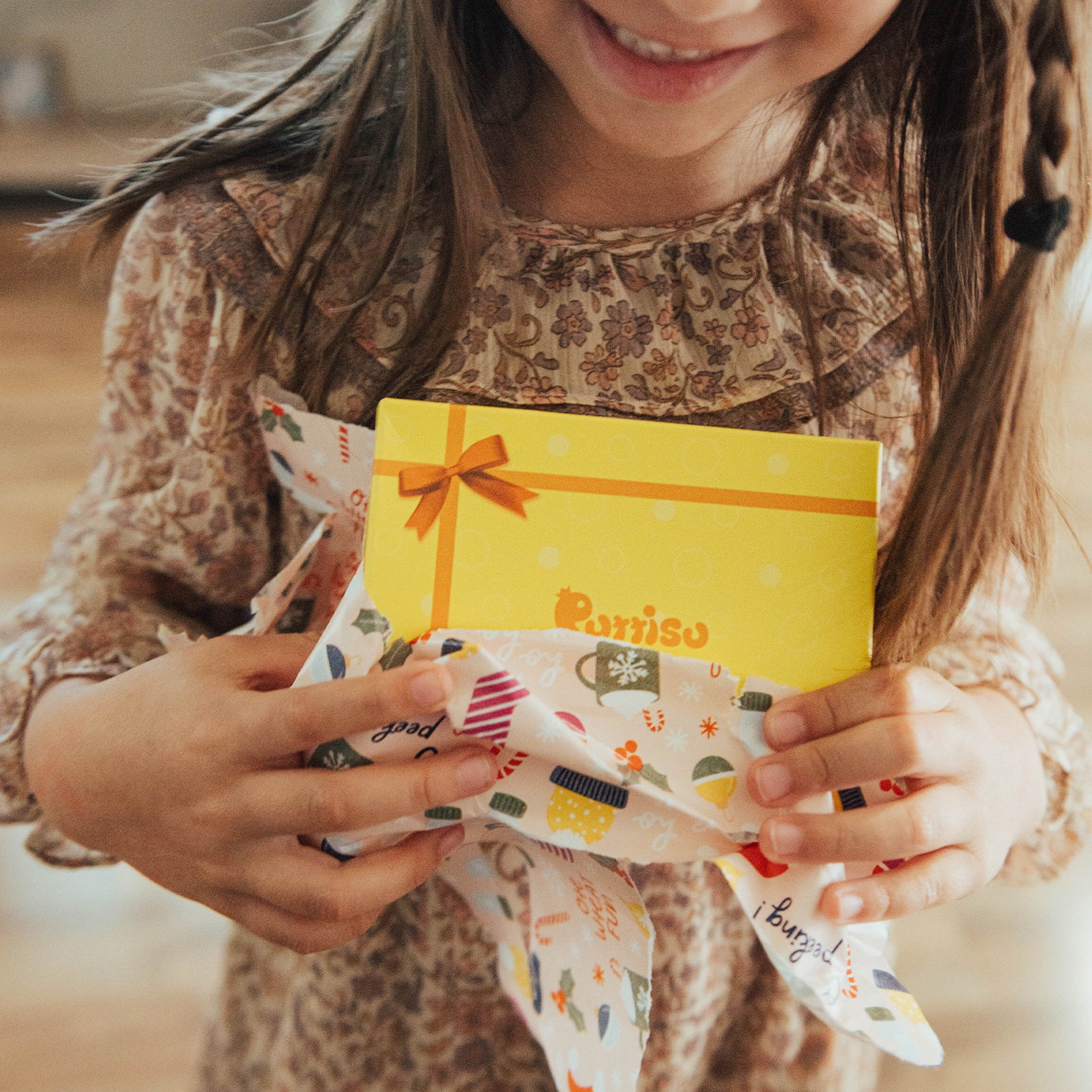 A girl smiling while unwrapping a yellow gift with a brown bow. She is wearing a floral dress.