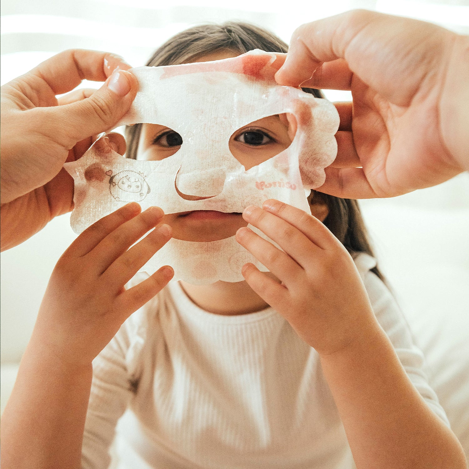 Girl wearing sheet face mask held by adult hands.