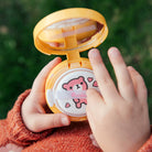 Child's hands holding an open, yellow, round compact of makeup on a green background. The compact has a bear design and hearts on the applicator. Child is wearing an orange sweater.