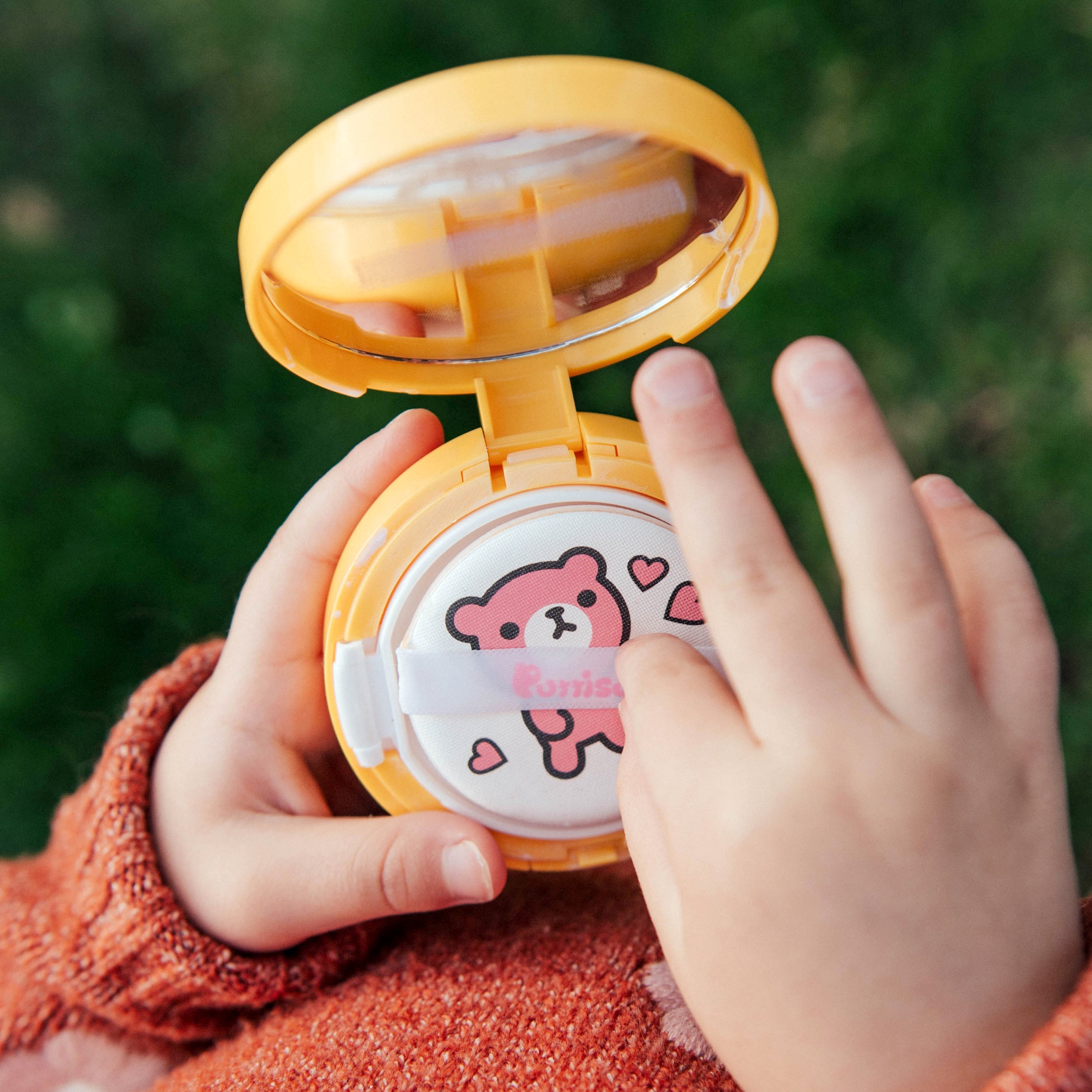 Child's hands holding an open, yellow, round compact of makeup on a green background. The compact has a bear design and hearts on the applicator. Child is wearing an orange sweater.