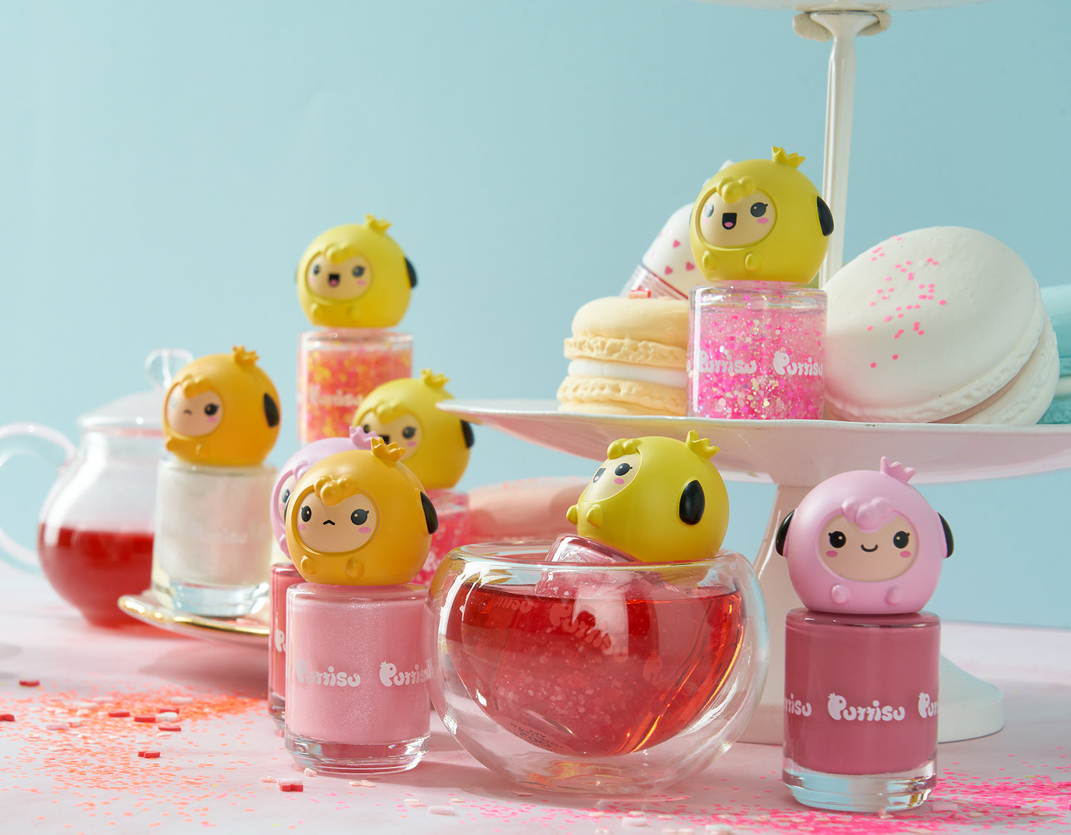 Close-up of colorful nail polish bottles with cartoon character tops, macarons on a tiered stand, and beverages on a light blue background with scattered confetti.