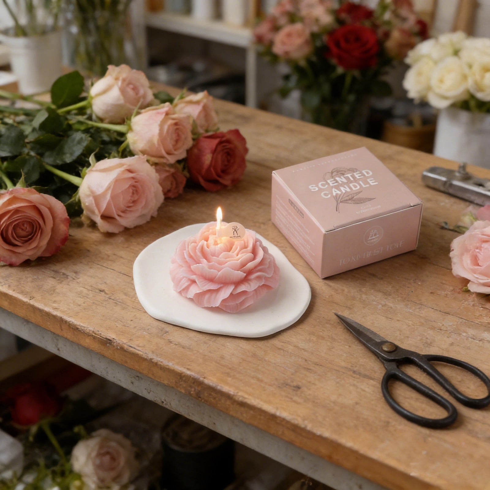 Pink peony flower soy wax candle by Jellycup, on a white ceramic dish near some roses and a candle box.
