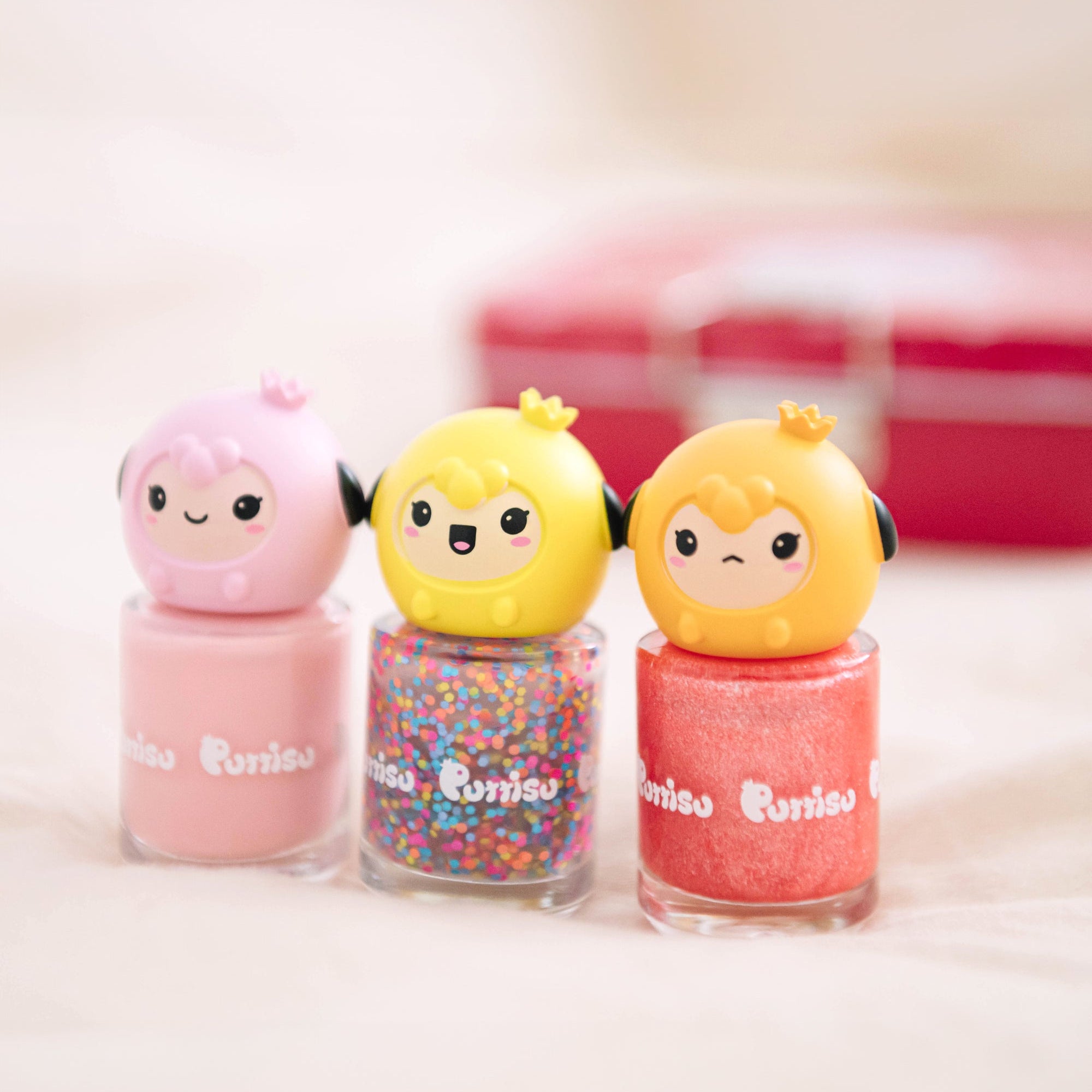 Three bottles of "Purriso" brand nail polish, each with a character-shaped cap. One is pink with a solid pink polish, one is yellow with confetti polish, and the other is orange with a textured polish. All three characters have black headphones. A red box is blurred in the background.