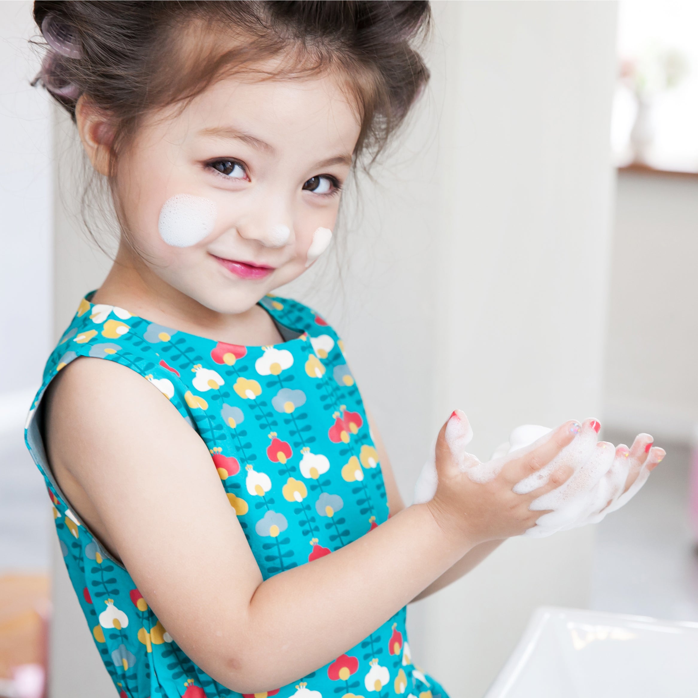 A young girl wearing a teal floral dress washes her hands with soap.