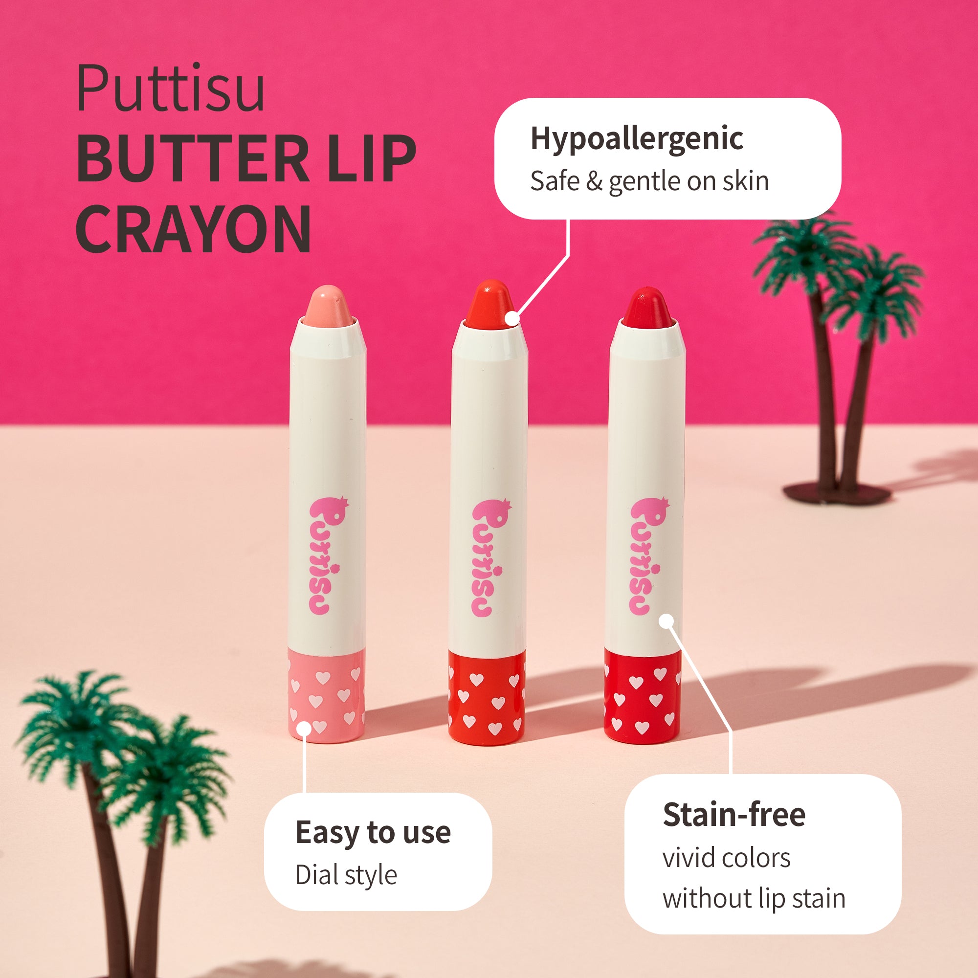 Three lip crayons in pink, red, and darker red with white bodies and heart-patterned bases, set against a pink and light pink background with miniature palm trees.
