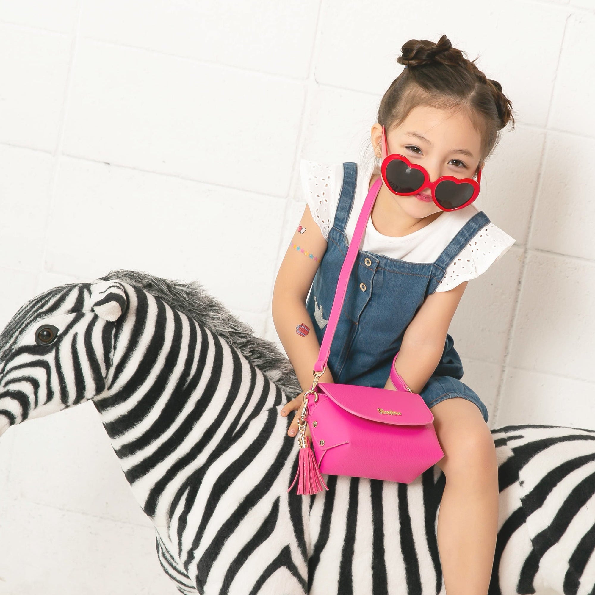 Child sitting on a zebra toy, wearing red heart sunglasses, denim dress, and a pink purse.