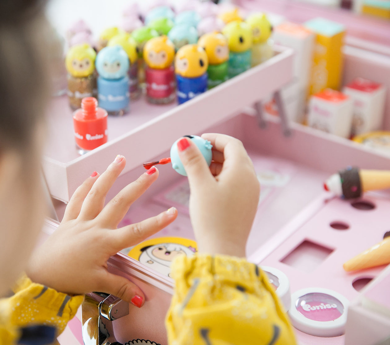 Child painting nails with red Puttisu kids nail polish from a character bottle, with a colorful nail polish collection displayed in a pink vanity case.