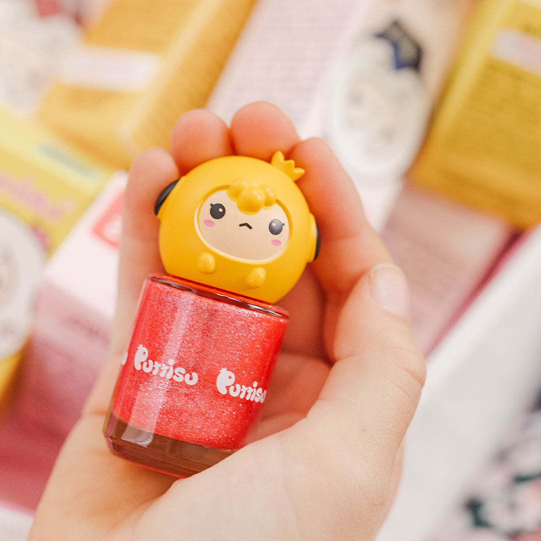 Close-up of a hand holding a red glitter nail polish with a yellow cartoon sheep head cap.