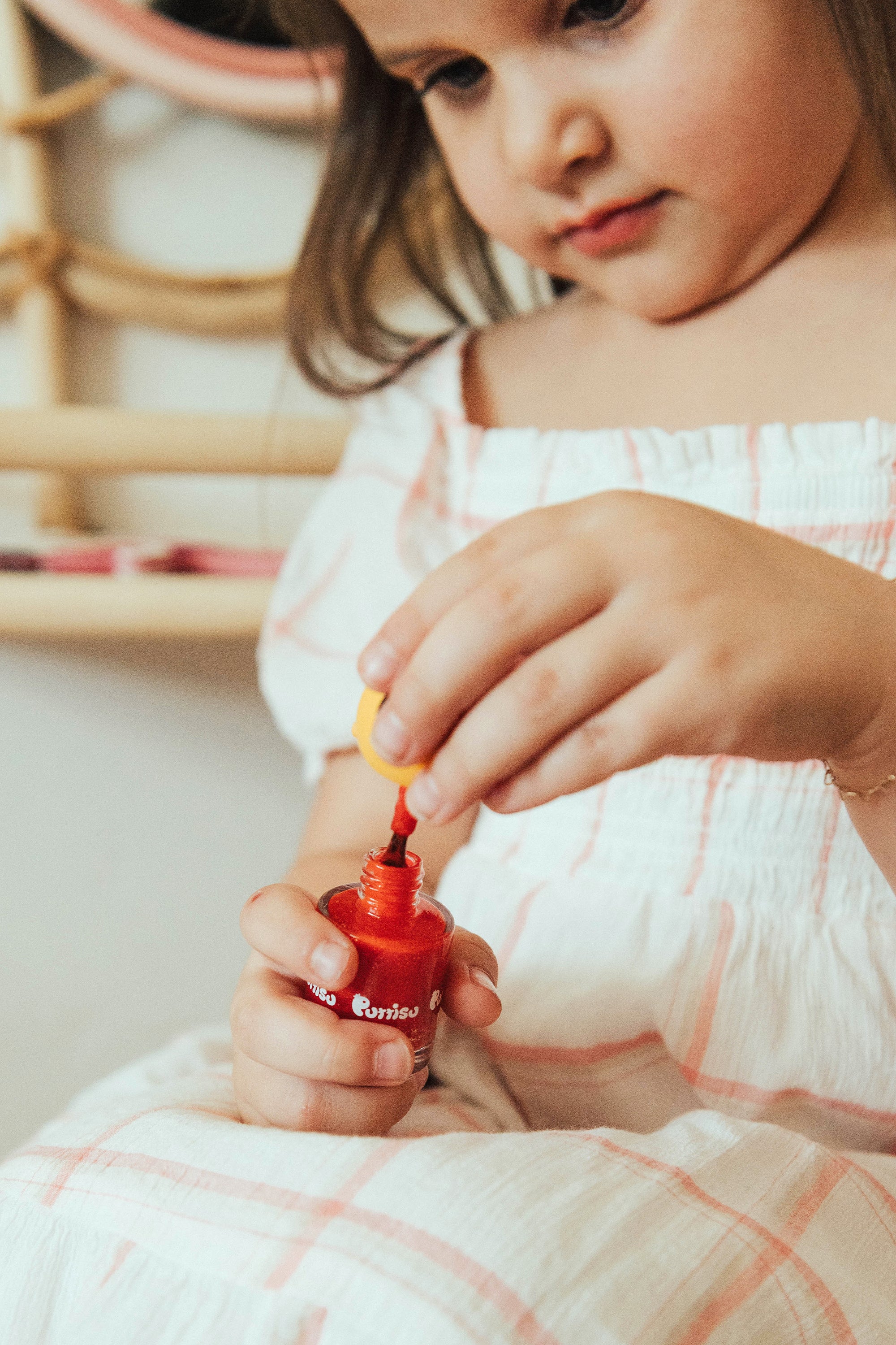 A young girl opening a bottle of red nail polish while wearing a white and pink plaid dress.