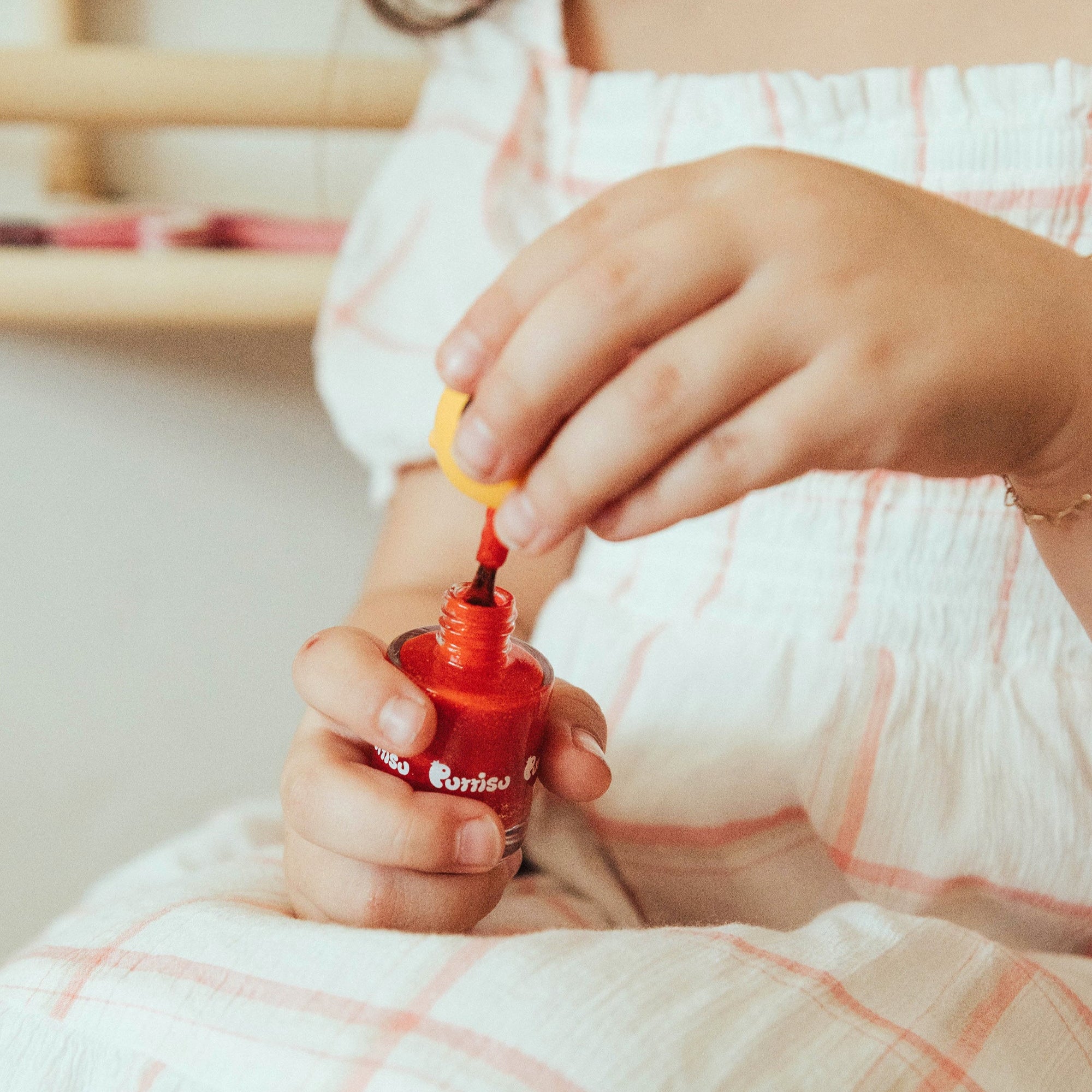 Young girl holding open bottle of red nail polish, wearing a white and red checked dress.