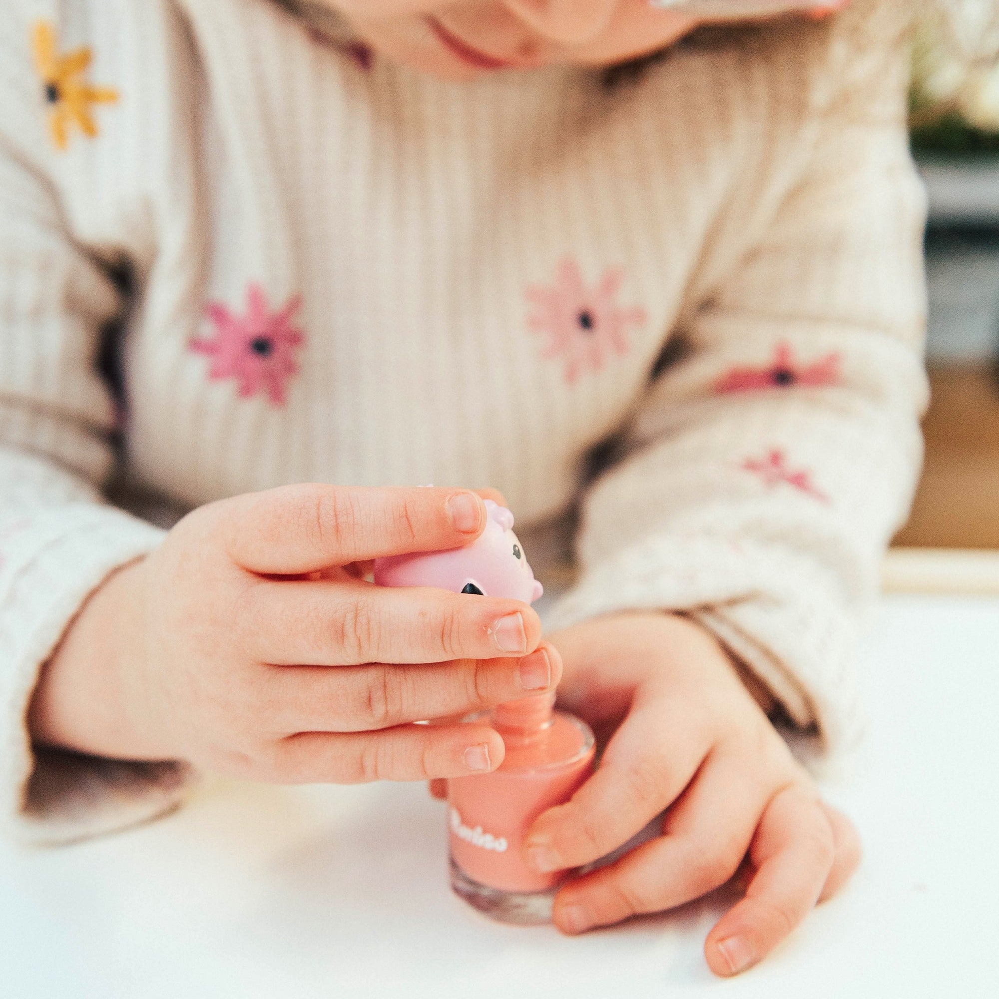 Child's hands holding pink nail polish, wearing flower-patterned knit sweater.