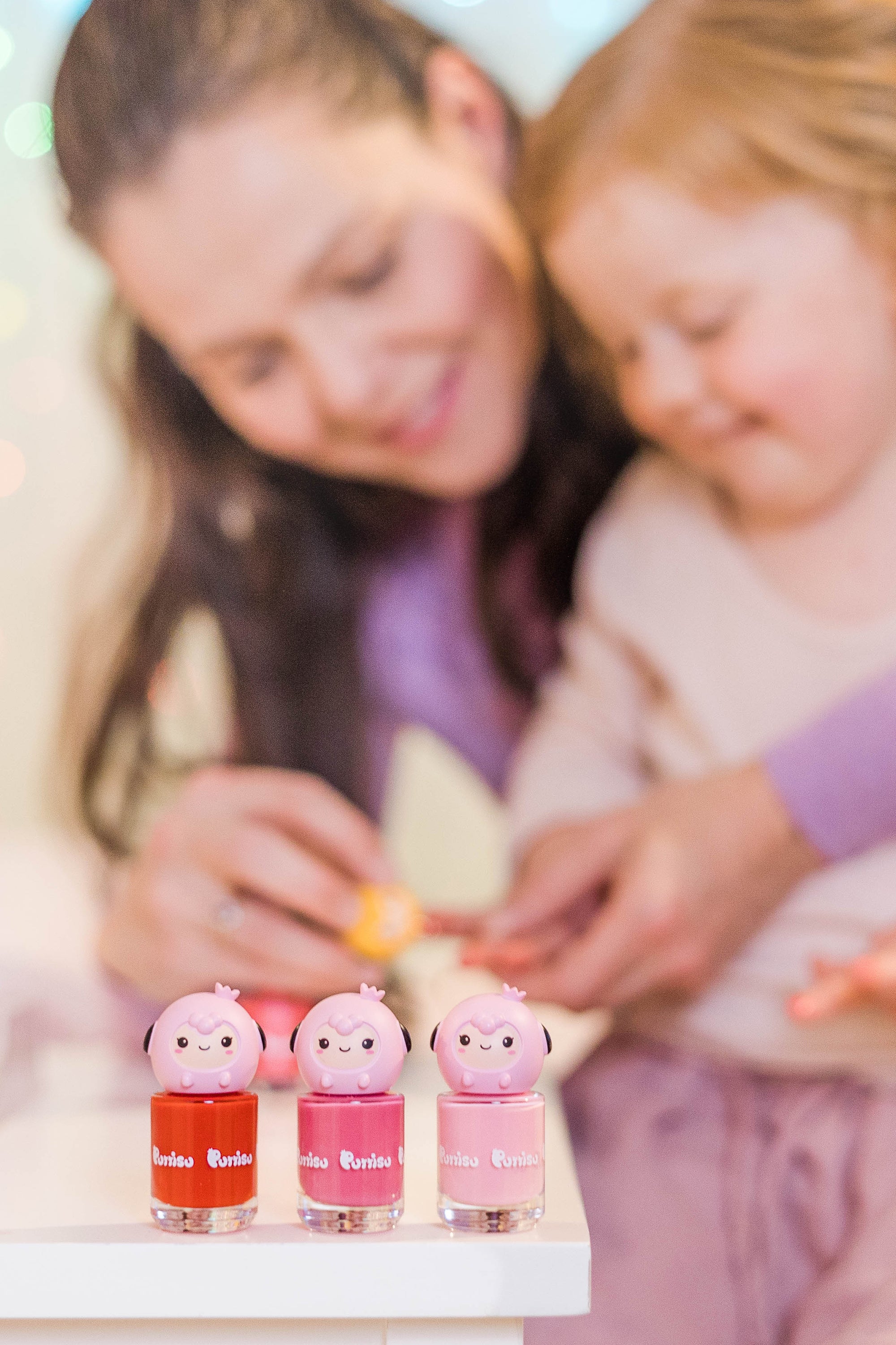 Three bottles of brightly colored nail polish with pink sheep-like character toppers. Mother and child in the blurred background.