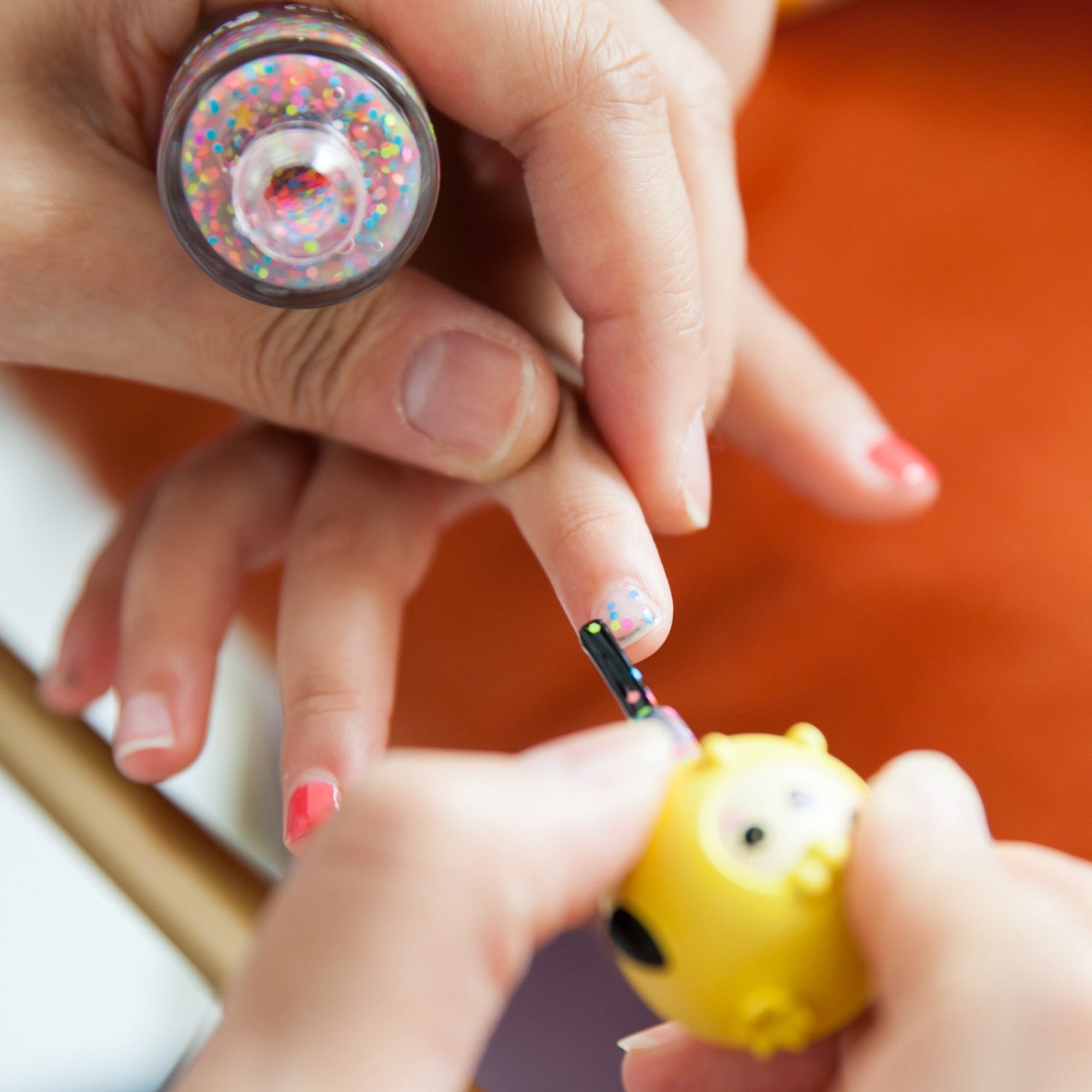 Close-up of nail polish application. Two bottles of nail polish; one with a yellow owl on the cap, and one with colorful glitter. Hand, nails.