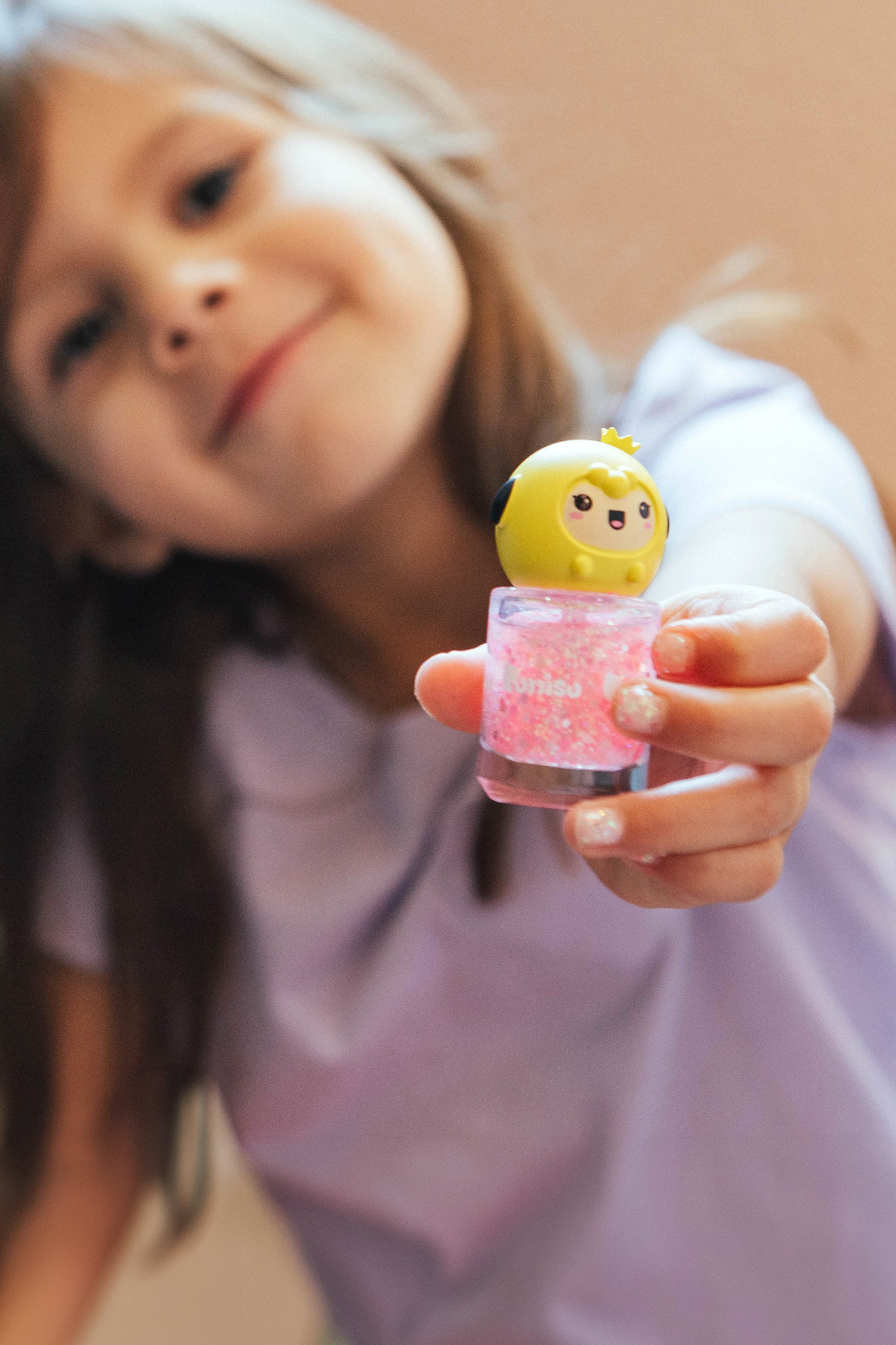 Girl holding pink glitter nail polish with a yellow cartoon cap.