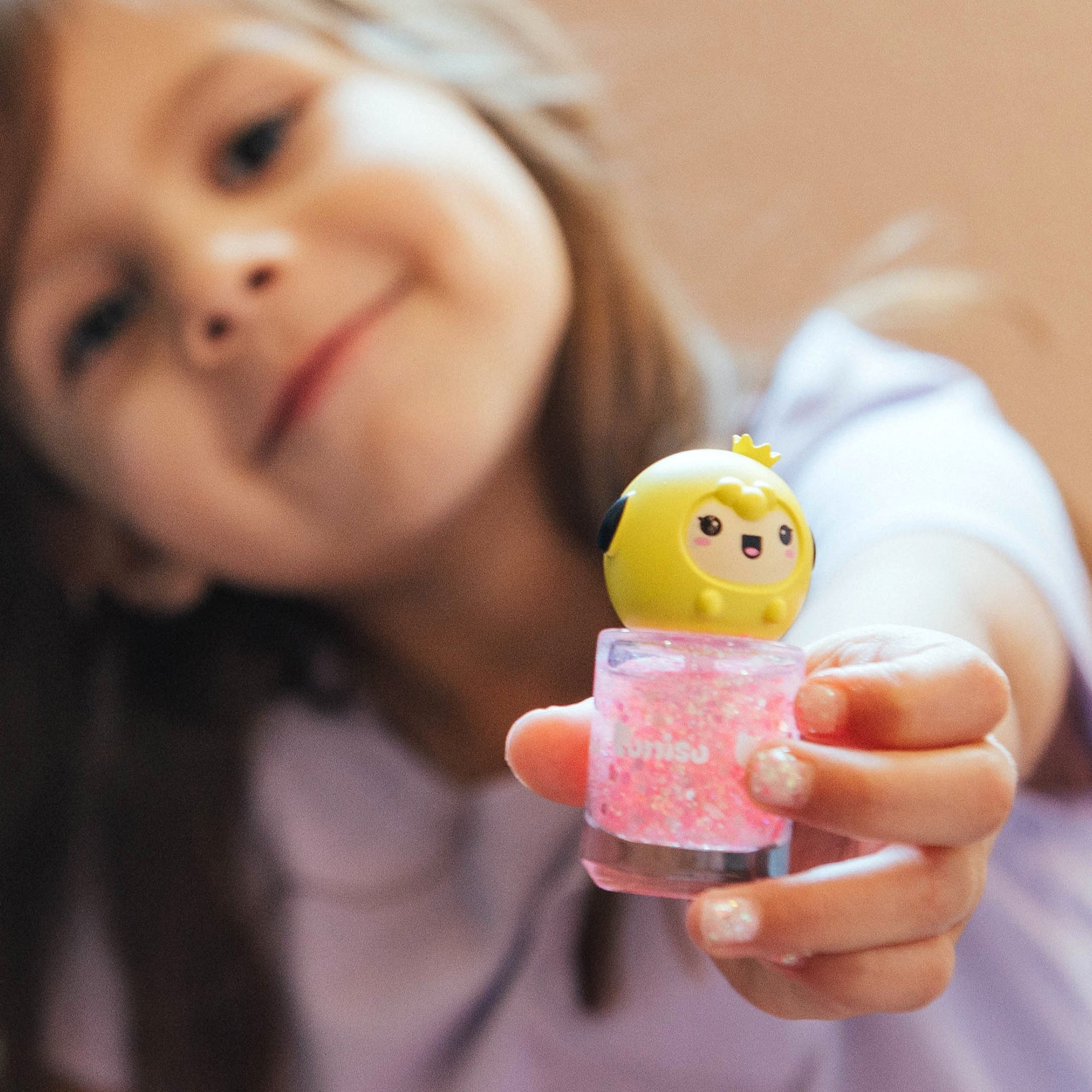 Smiling child holds pink glitter nail polish with a yellow cartoon character cap.