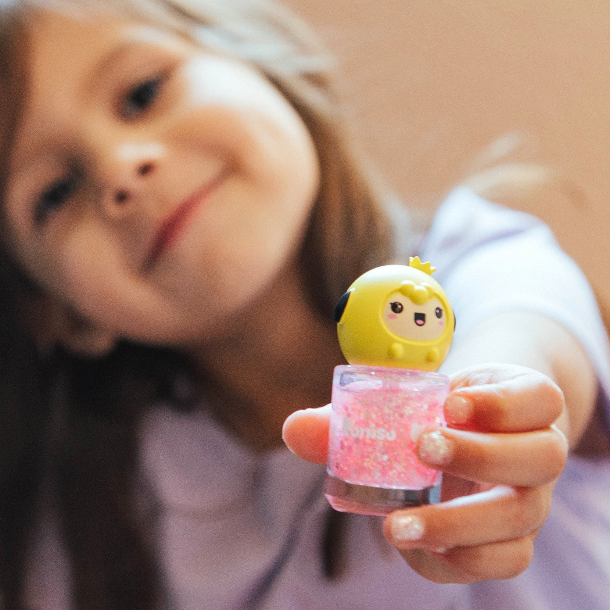 A smiling girl holds up pink glitter nail polish with a yellow cartoon animal cap.