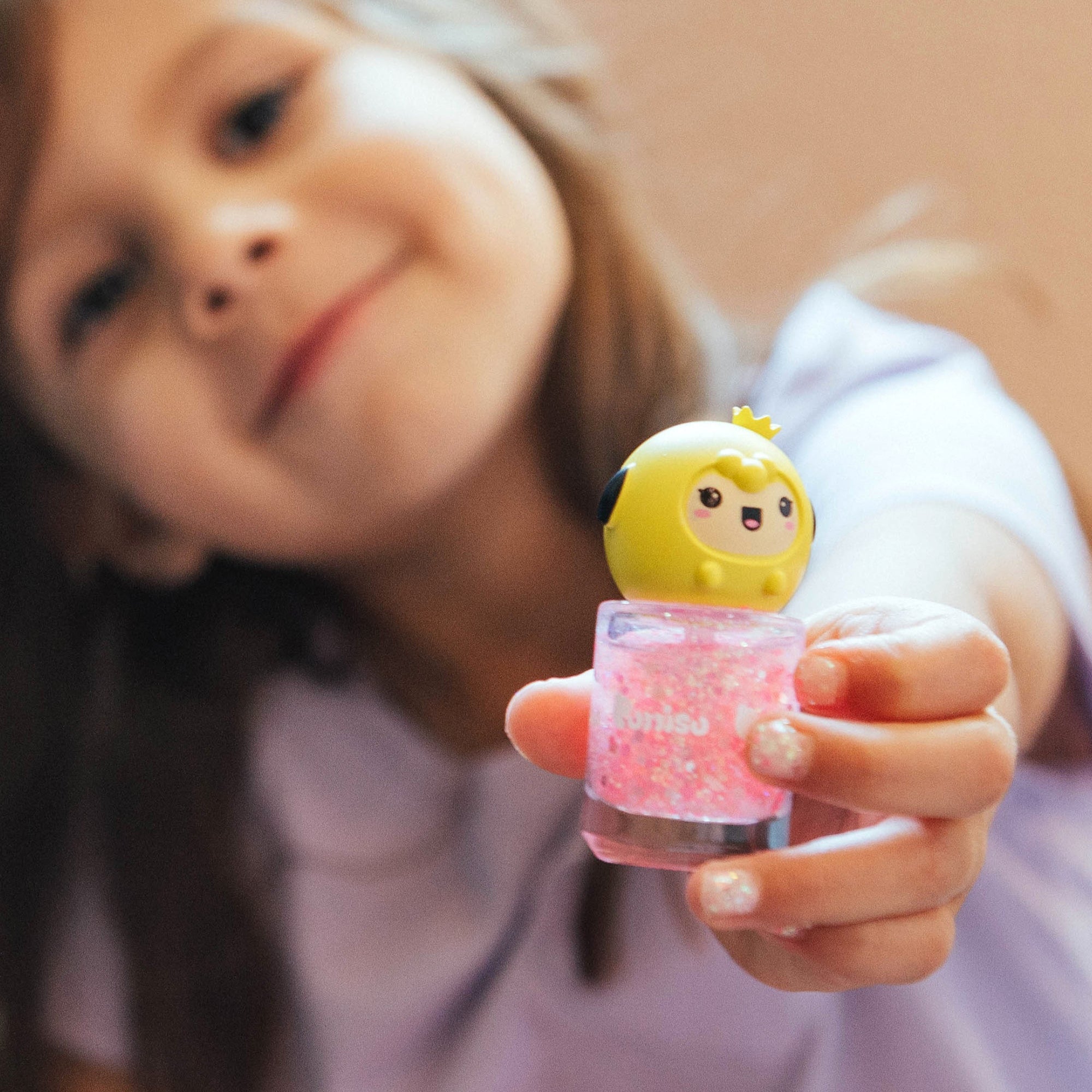 A young girl with light brown hair holds a bottle of pink glitter nail polish topped with a yellow cartoon character.