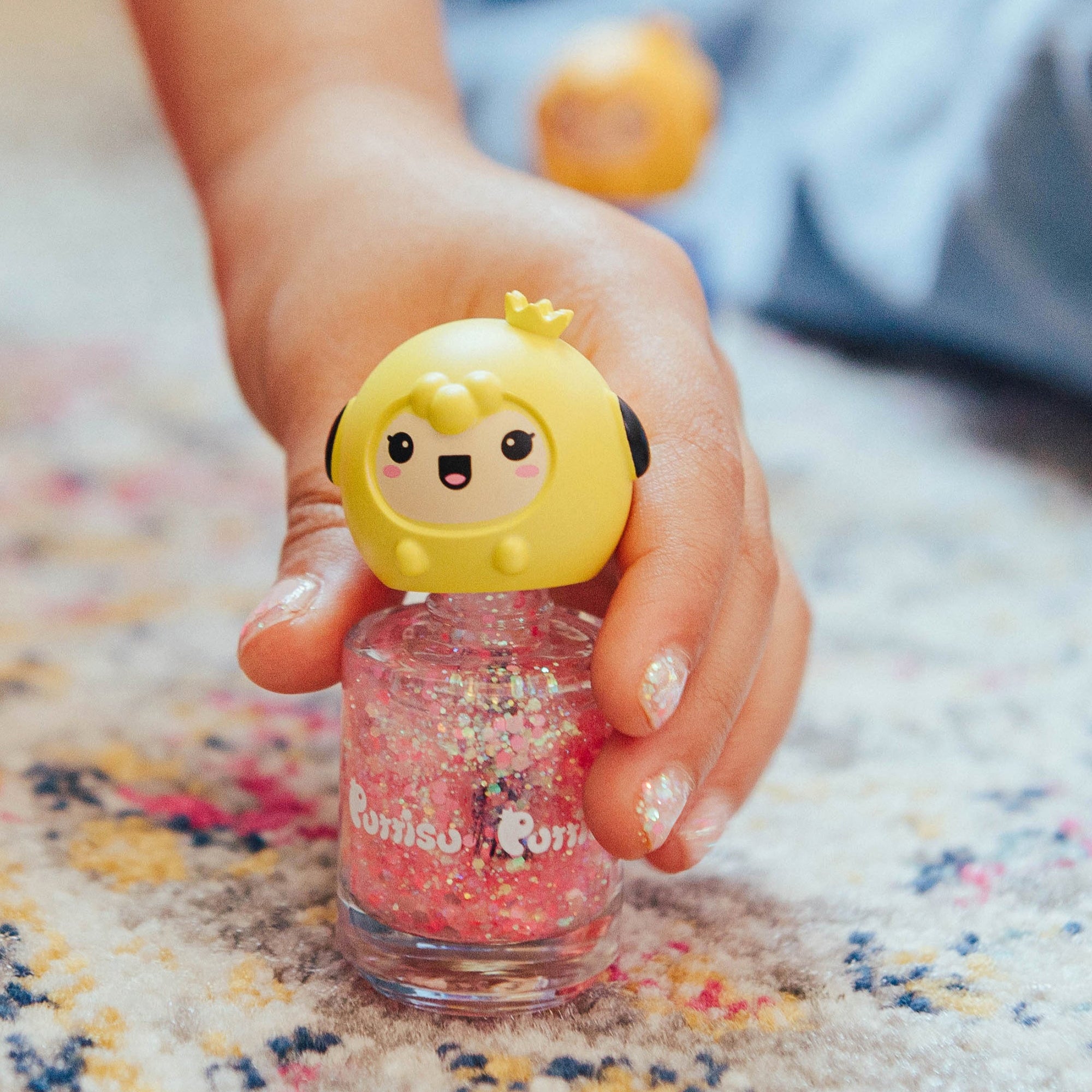 Hand holding pink glitter nail polish with a yellow cartoon character topper on a patterned rug.