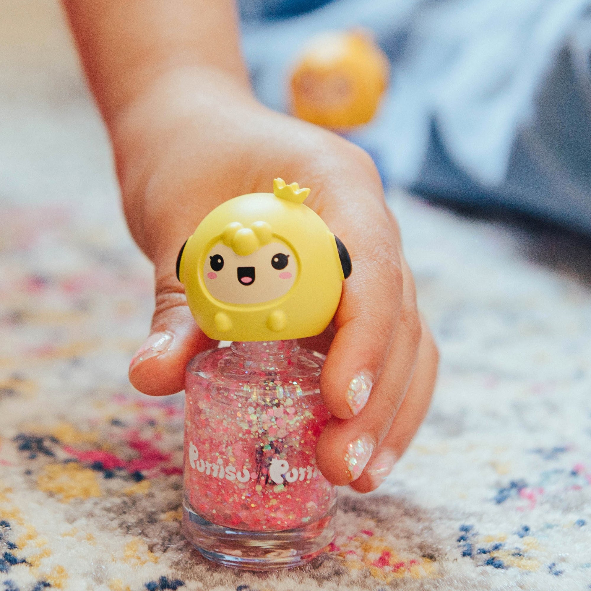 Hand holding a pink glitter nail polish with yellow cartoon cap.