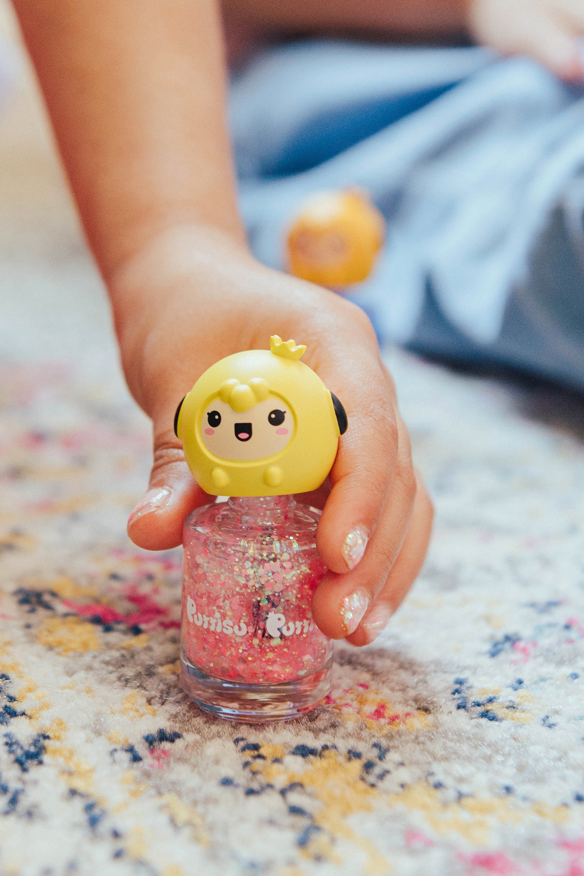Child's hand holding glittery pink nail polish with a yellow cartoon cap.