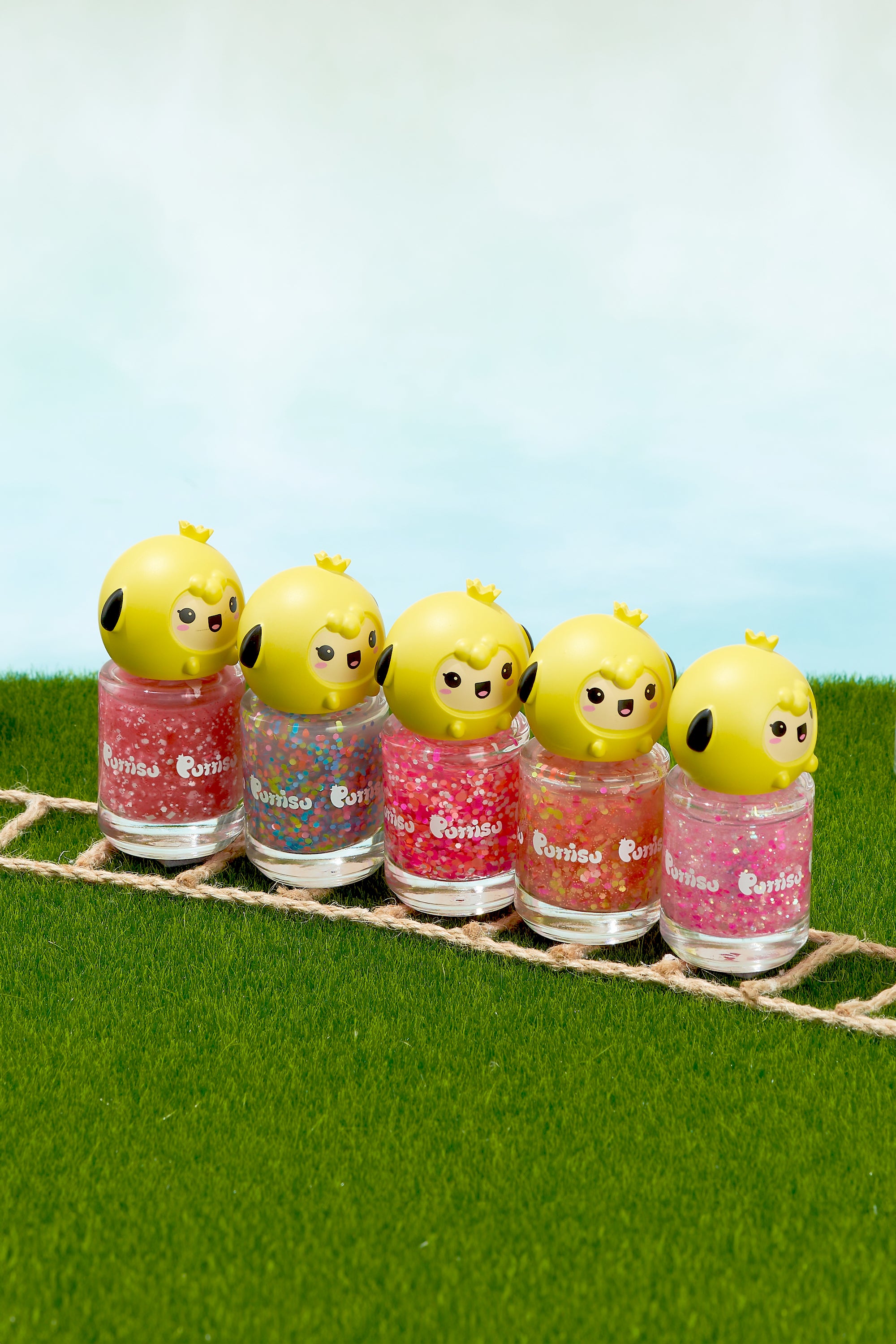 Five bottles of colorful glitter nail polish with cartoon chick tops sit on a green surface with a sky backdrop.