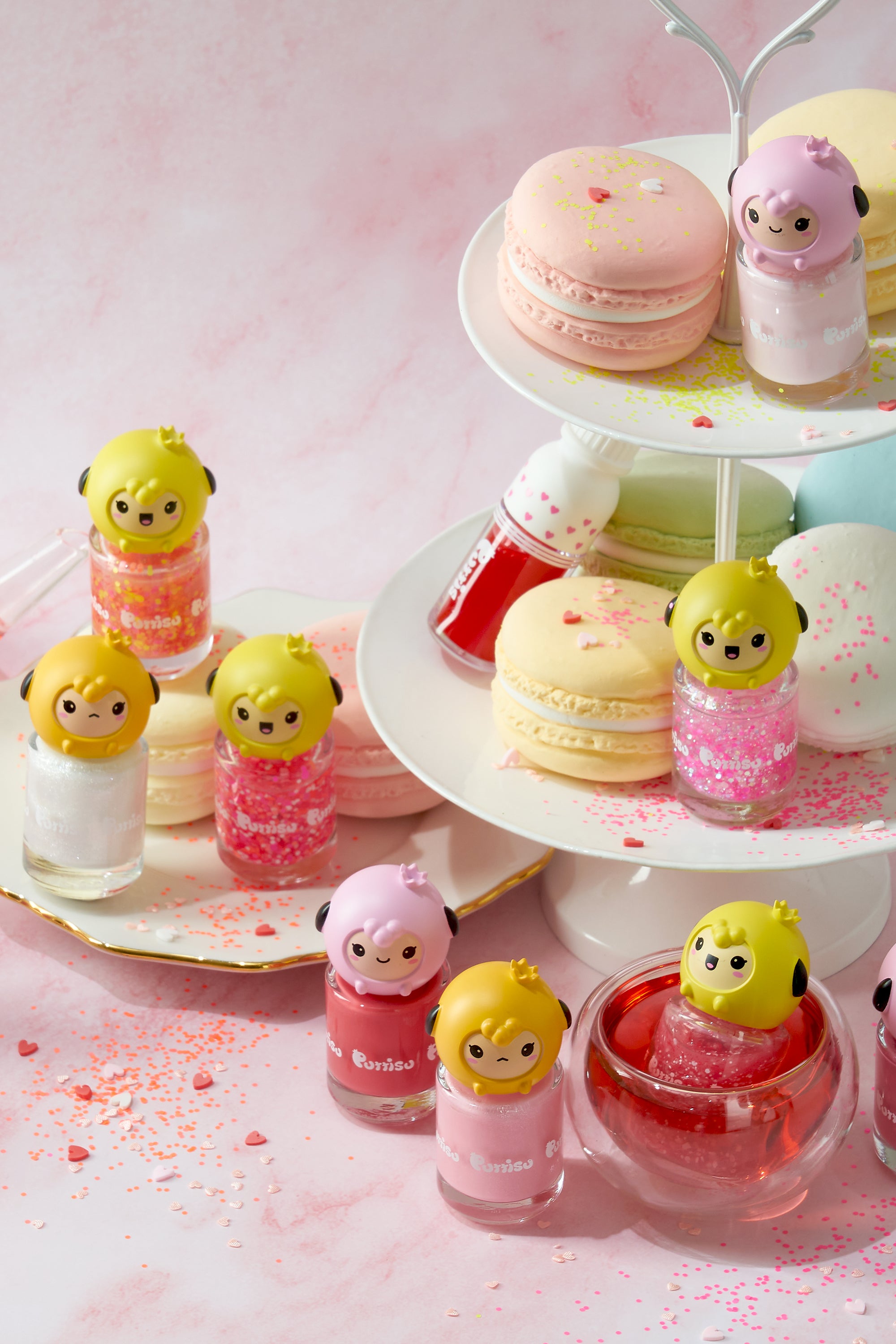 A three-tiered dessert stand with pastel macarons and nail polish bottles topped with cartoon lamb figures. The backdrop is a pink marbled surface scattered with sprinkles.
