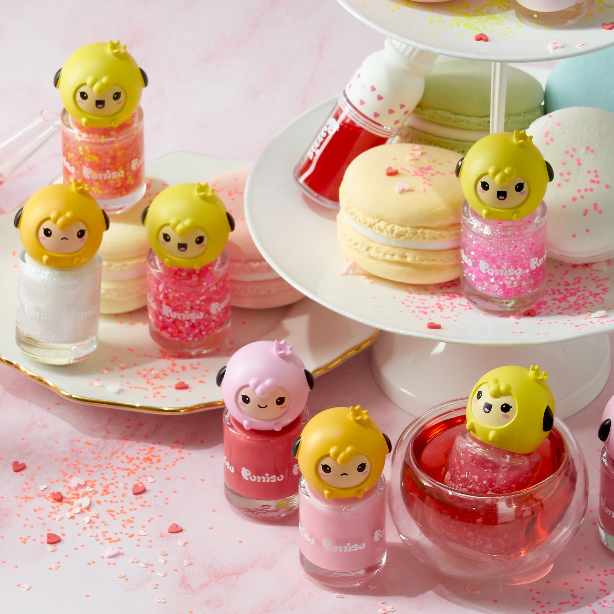 Several bottles of colorful nail polish with cartoon sheep-like figures on top are displayed on a pink surface and a tiered cake stand, surrounded by macarons and scattered heart-shaped sprinkles.