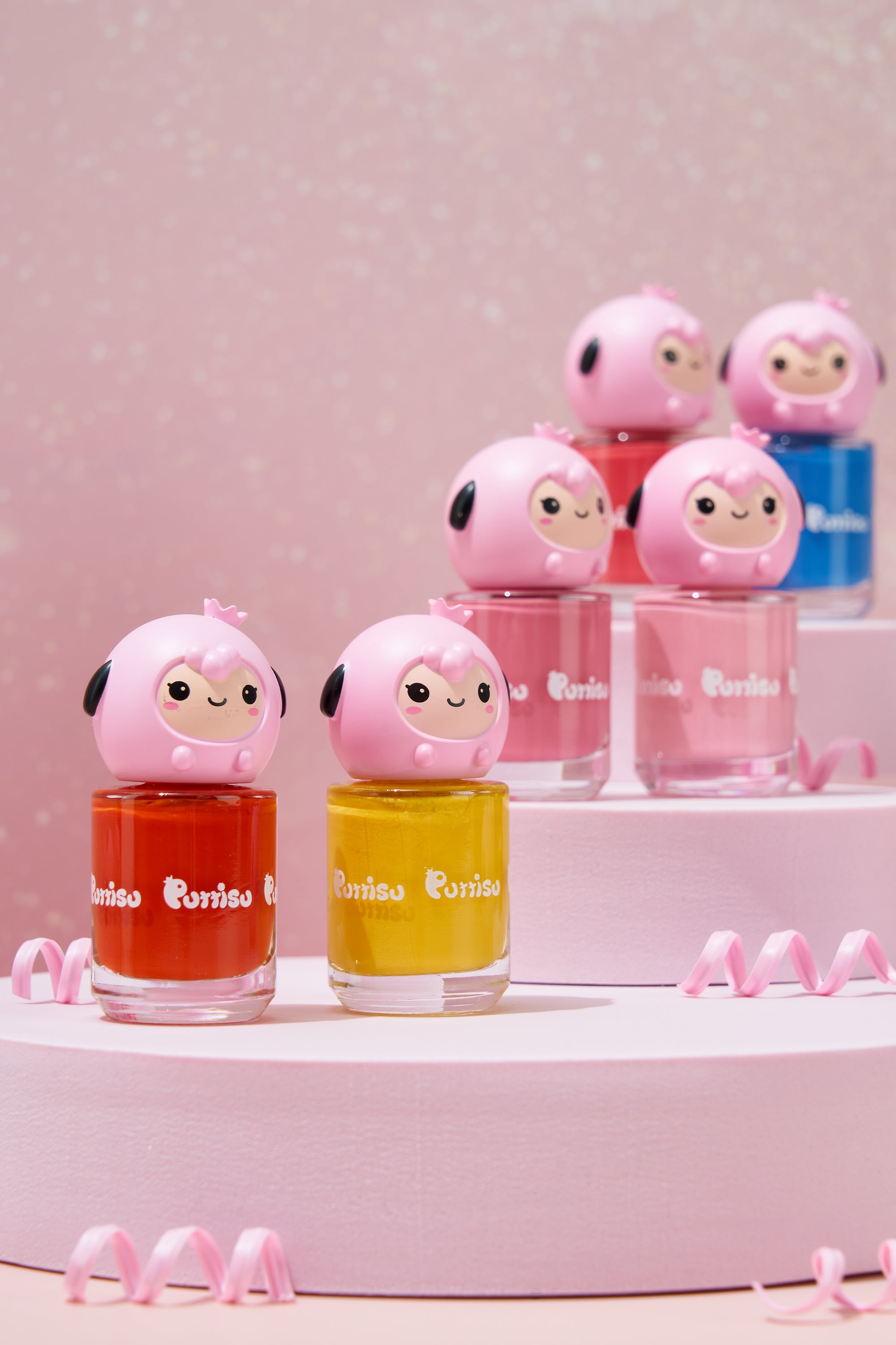 Several bottles of colorful nail polish with pink, cartoonish sheep-shaped lids arranged on a multi-tiered pink platform, accompanied by pink curly ribbons.