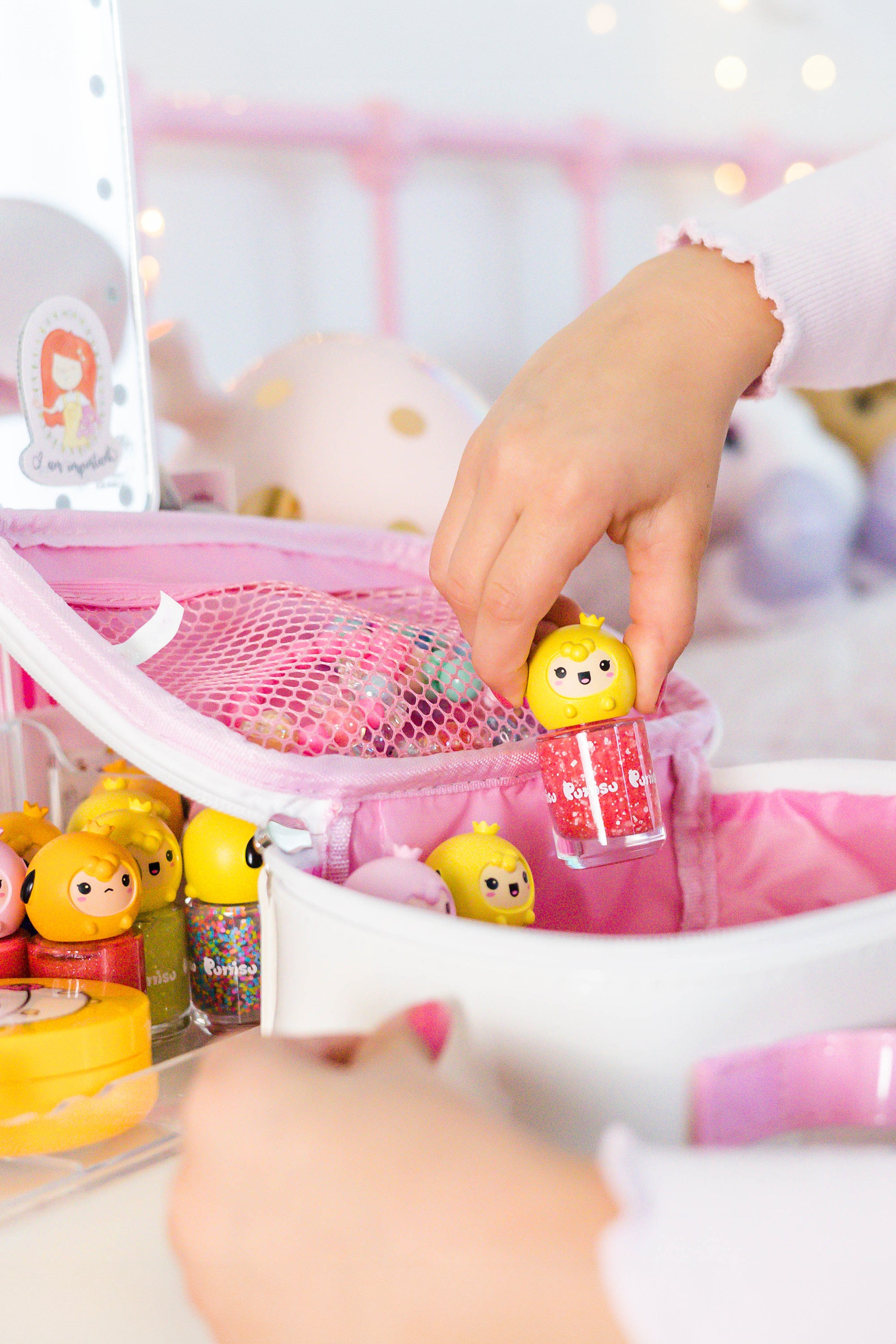 A child's hand holding a yellow and red "Purr-iso" nail polish above a pink cosmetic bag filled with similar nail polishes.