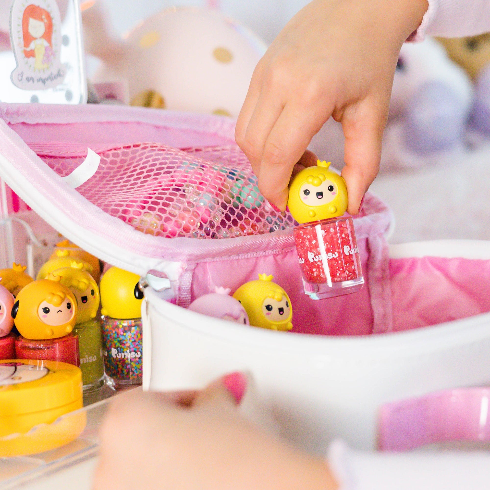 Child's hand holding red glitter nail polish with yellow character cap above open white and pink makeup bag with cosmetics.