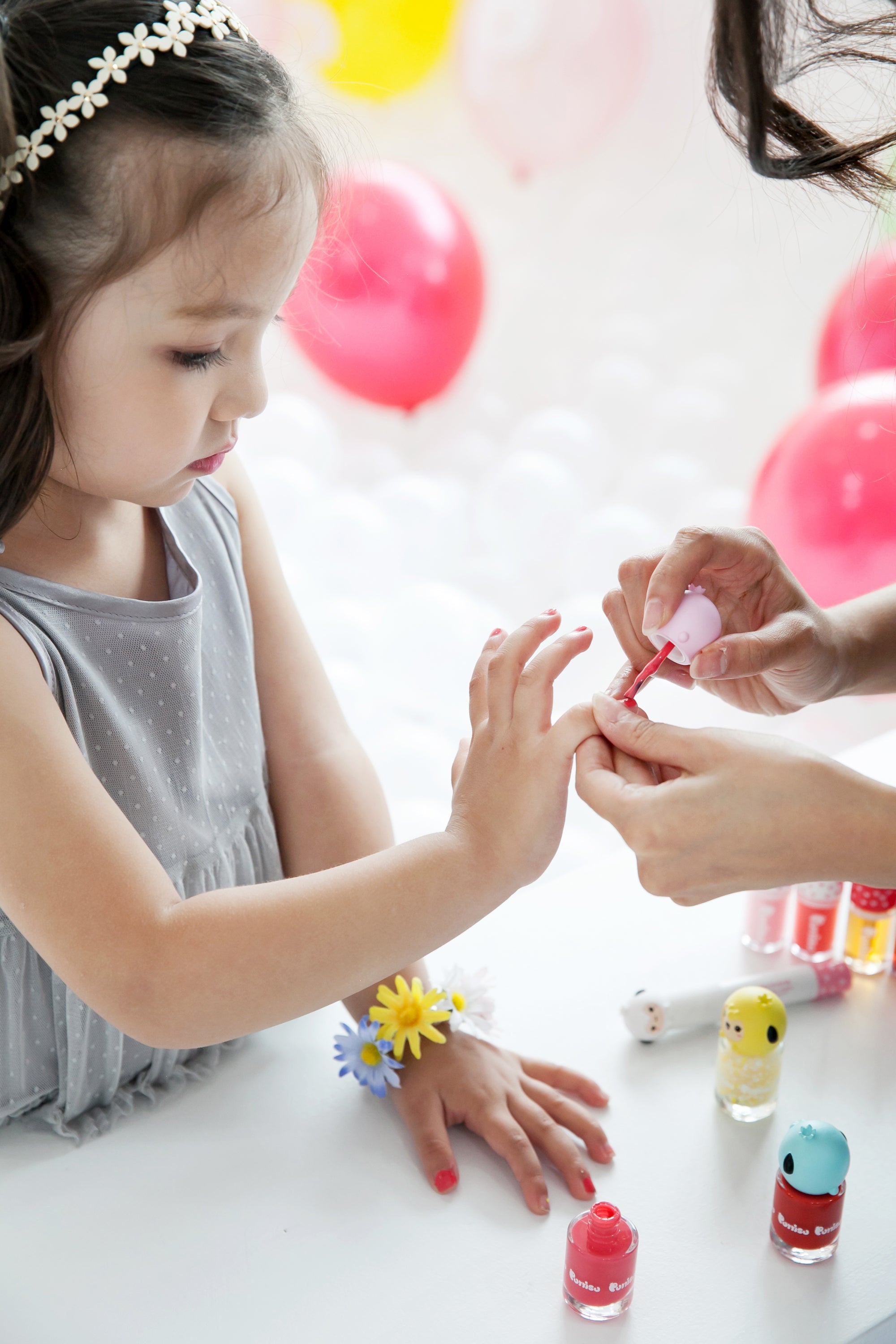 A little girl getting her nails painted red, wearing a gray dress and a daisy headband.