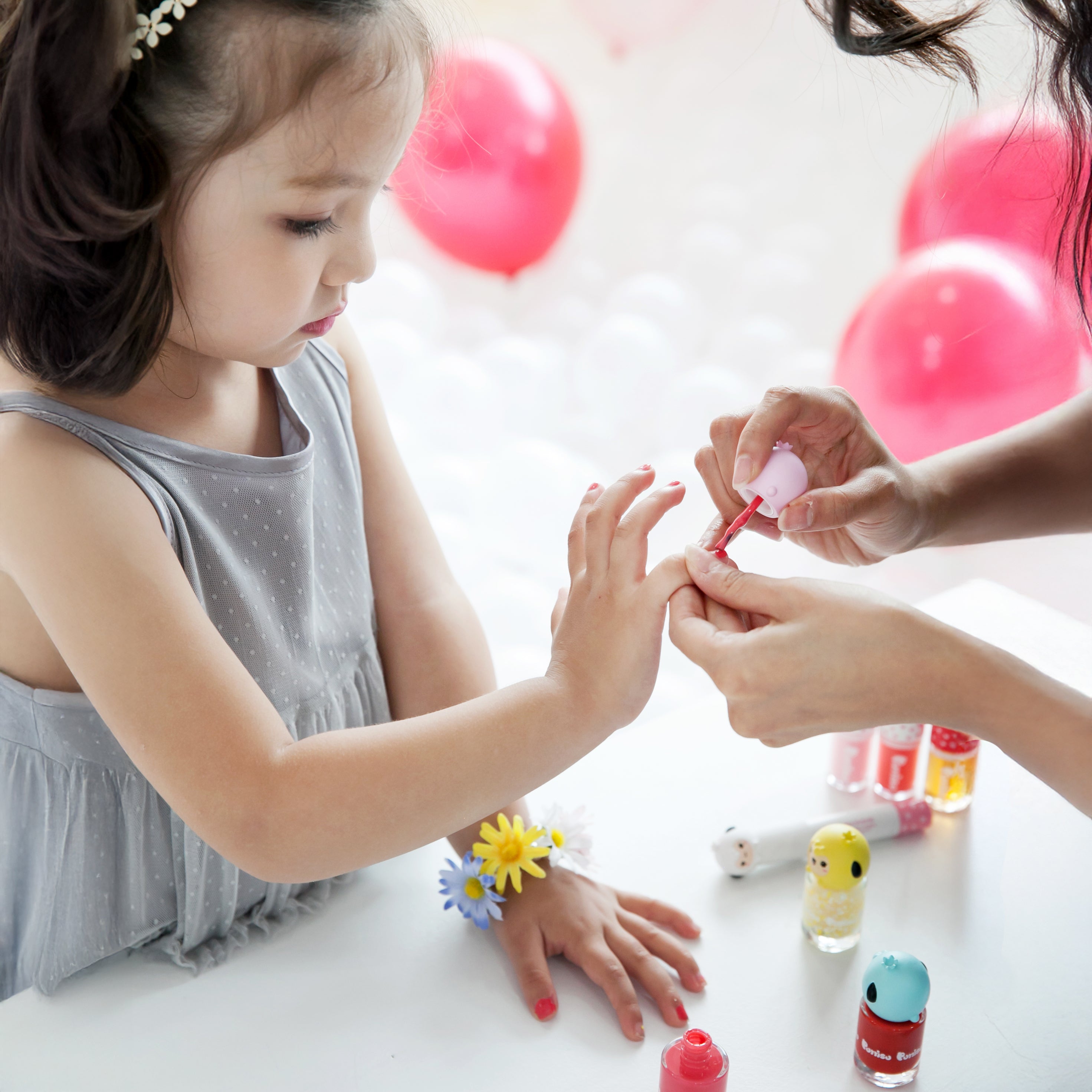 Child in gray dress with flower bracelet getting red nail polish applied; colorful polish bottles and balloons in background.