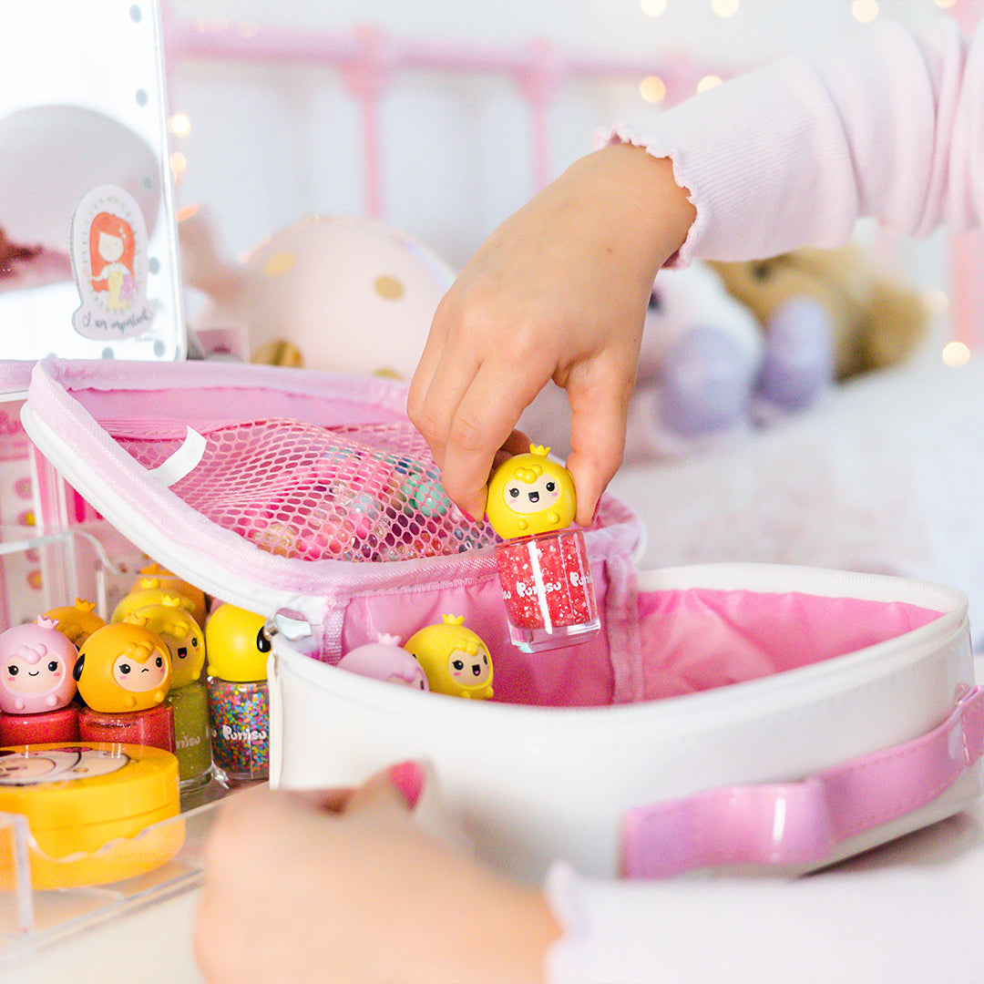 Child's hand holding yellow bird-shaped nail polish top over red glitter polish bottle, pink and white makeup bag, desk.