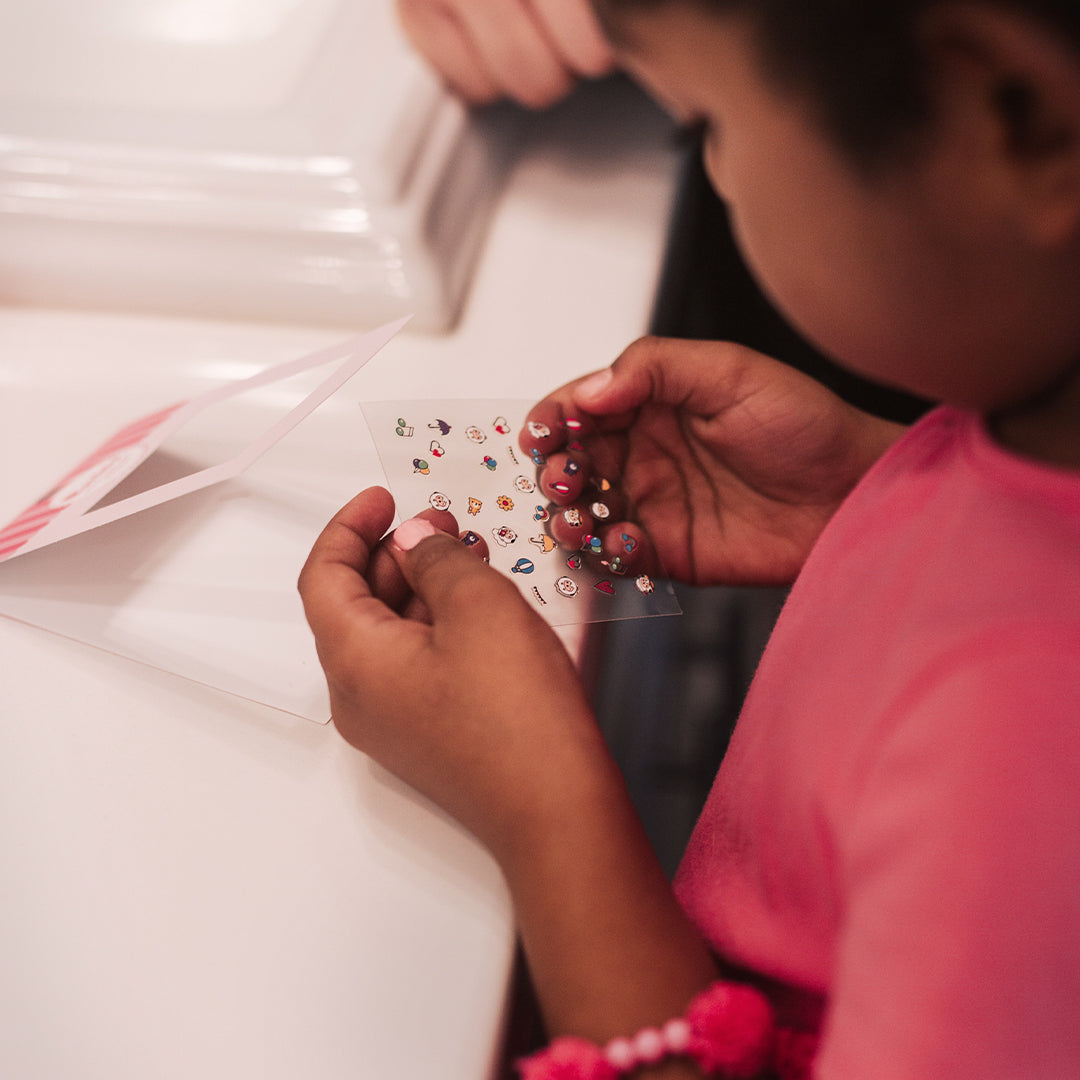 A child with dark skin wearing a pink shirt and a beaded bracelet holding a sheet of small, colorful stickers in front of an open card on a white table.