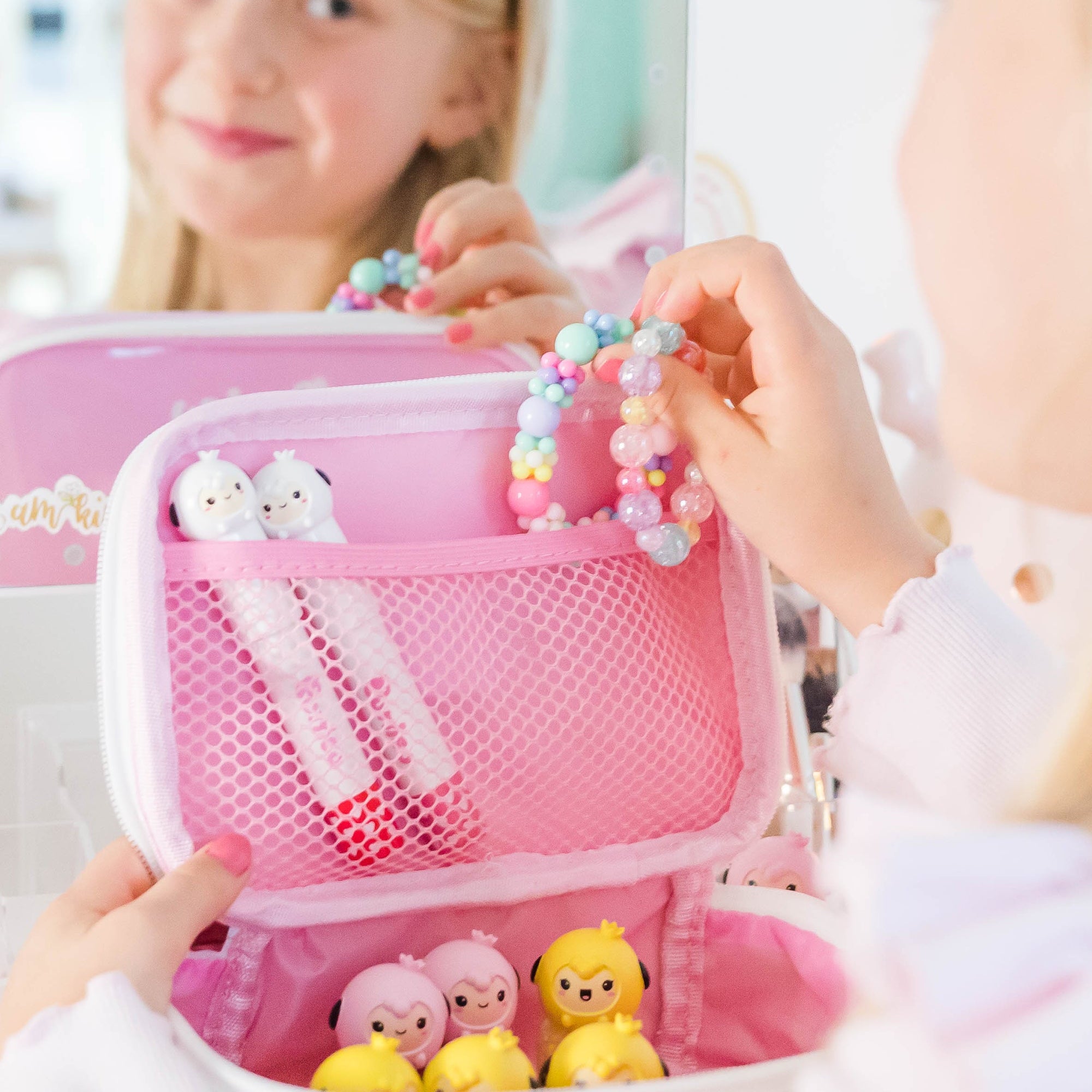 Young girl holding a beaded bracelet with a pink makeup case in front of a mirror.
