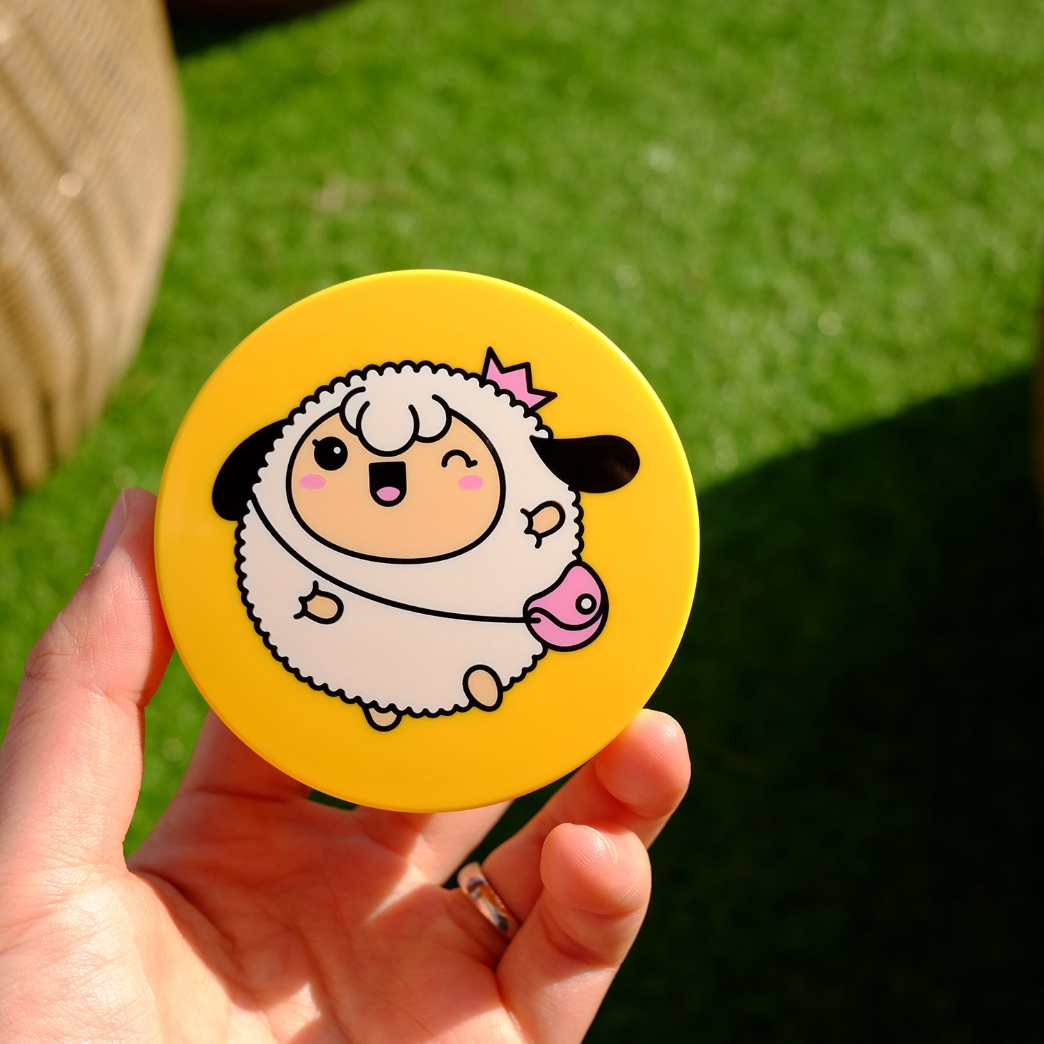 Hand holding a yellow round compact with a cartoon sheep wearing a crown.
