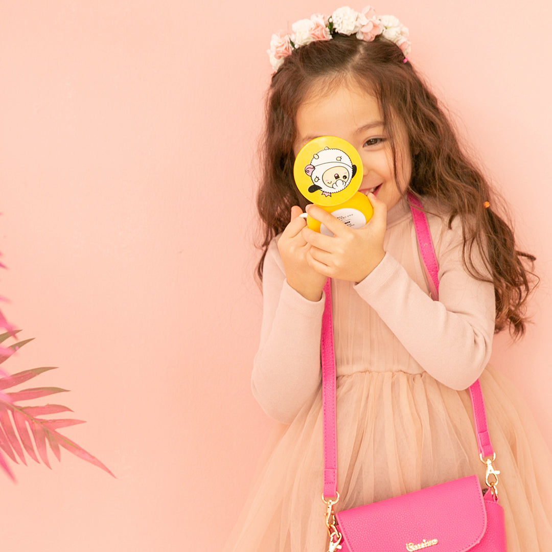 Girl in a peach dress and floral headband, covering her eye with a yellow compact, with a pink purse.