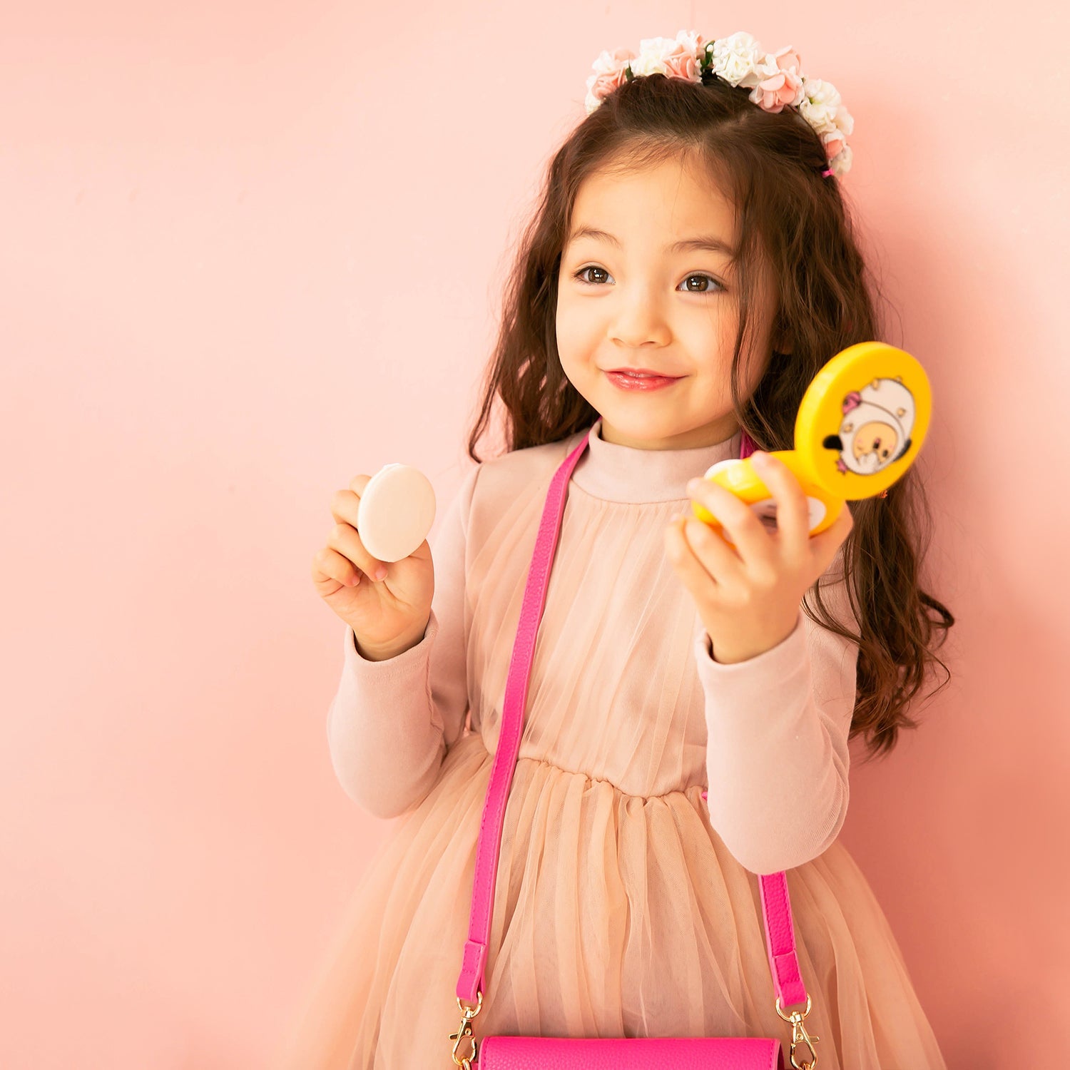A young girl with long hair and a flower crown holds makeup compacts, wearing a pink dress and purse in front of a pink backdrop.