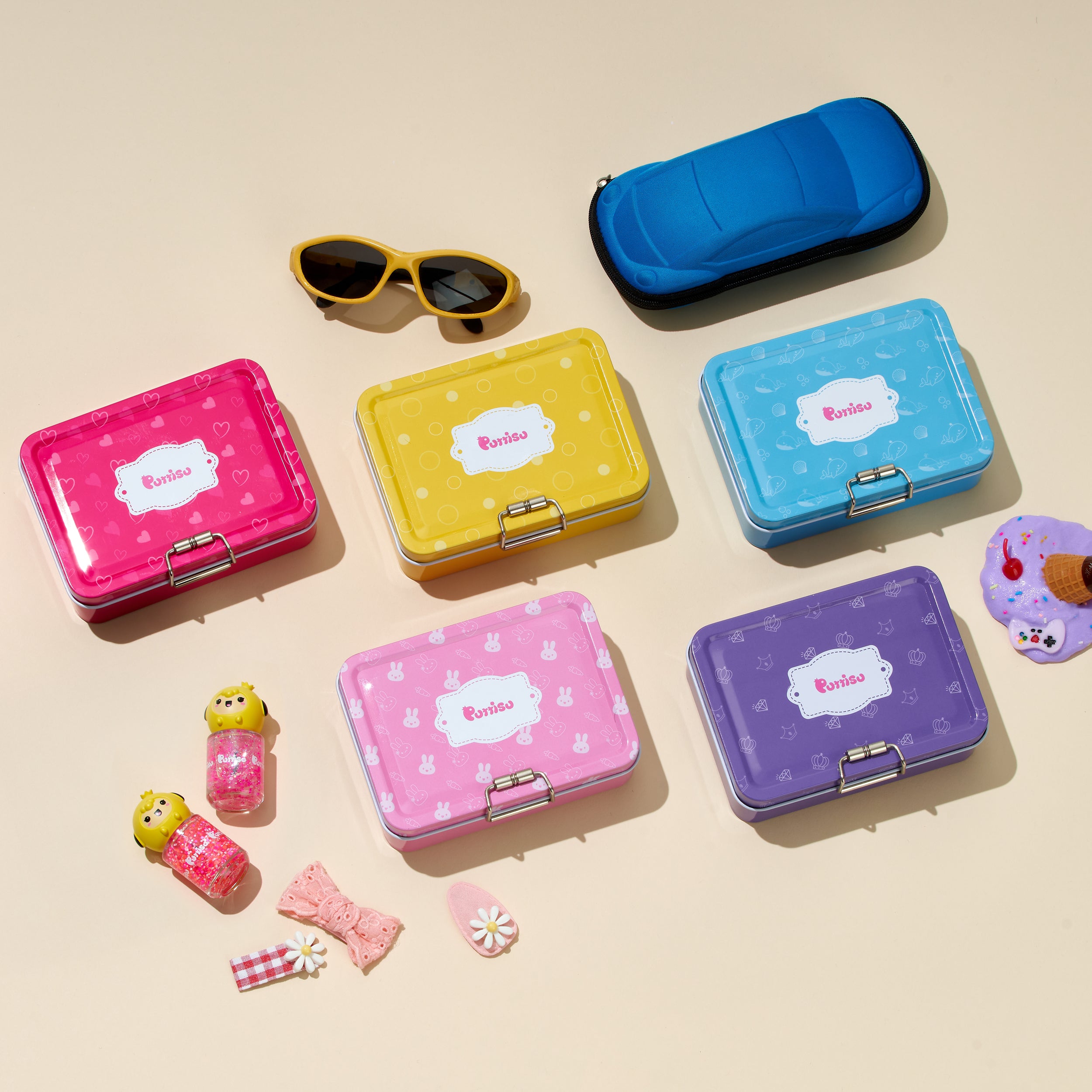 Group of colorful "Purrisu" lunchboxes, nail polish, sunglasses, hair accessories, and a blue car-shaped case on a neutral surface.