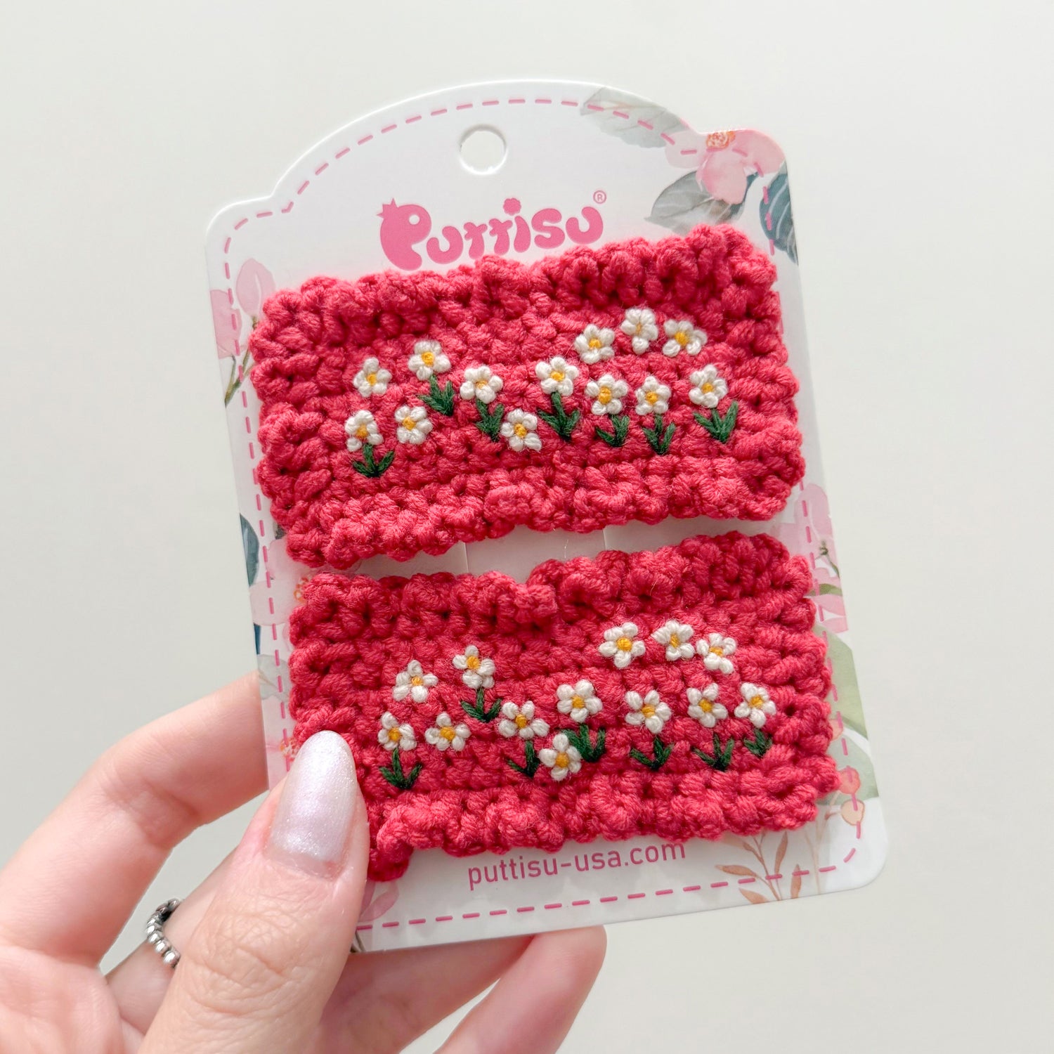 Two red crochet hair clips with embroidered white daisies on a floral-patterned card held in a hand.