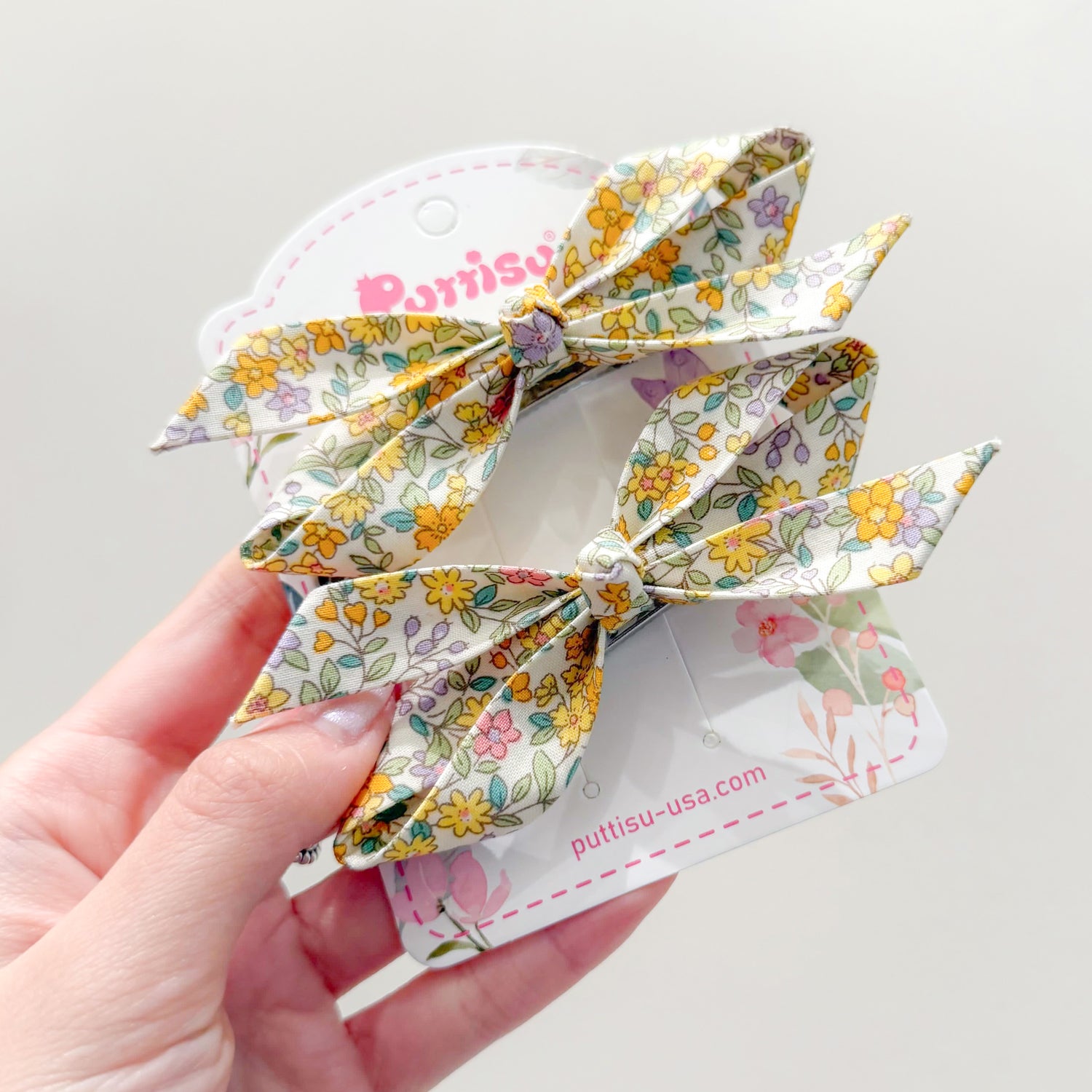 Two floral hair bows held in a hand. The bows have a white background with yellow, pink, and purple flowers. The bows are attached to a white cardboard tag.