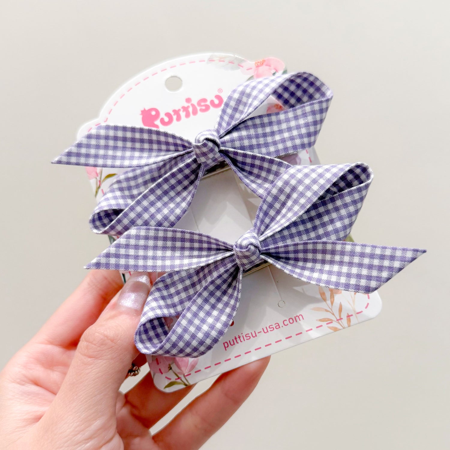 Two purple and white gingham hair clips on a floral-decorated card being held in a hand.