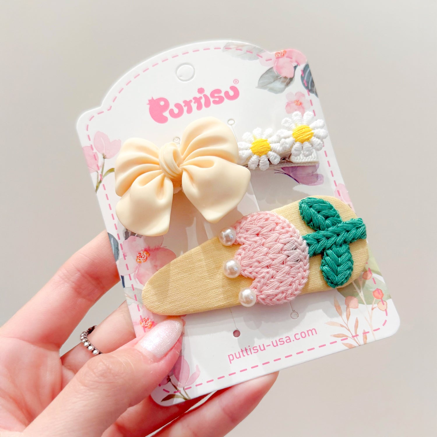 Three hair clips on a floral-patterned card. Cream-colored bow, two white daisy clips, and an oblong clip with pink crochet, pearls, and a green crochet leaf.