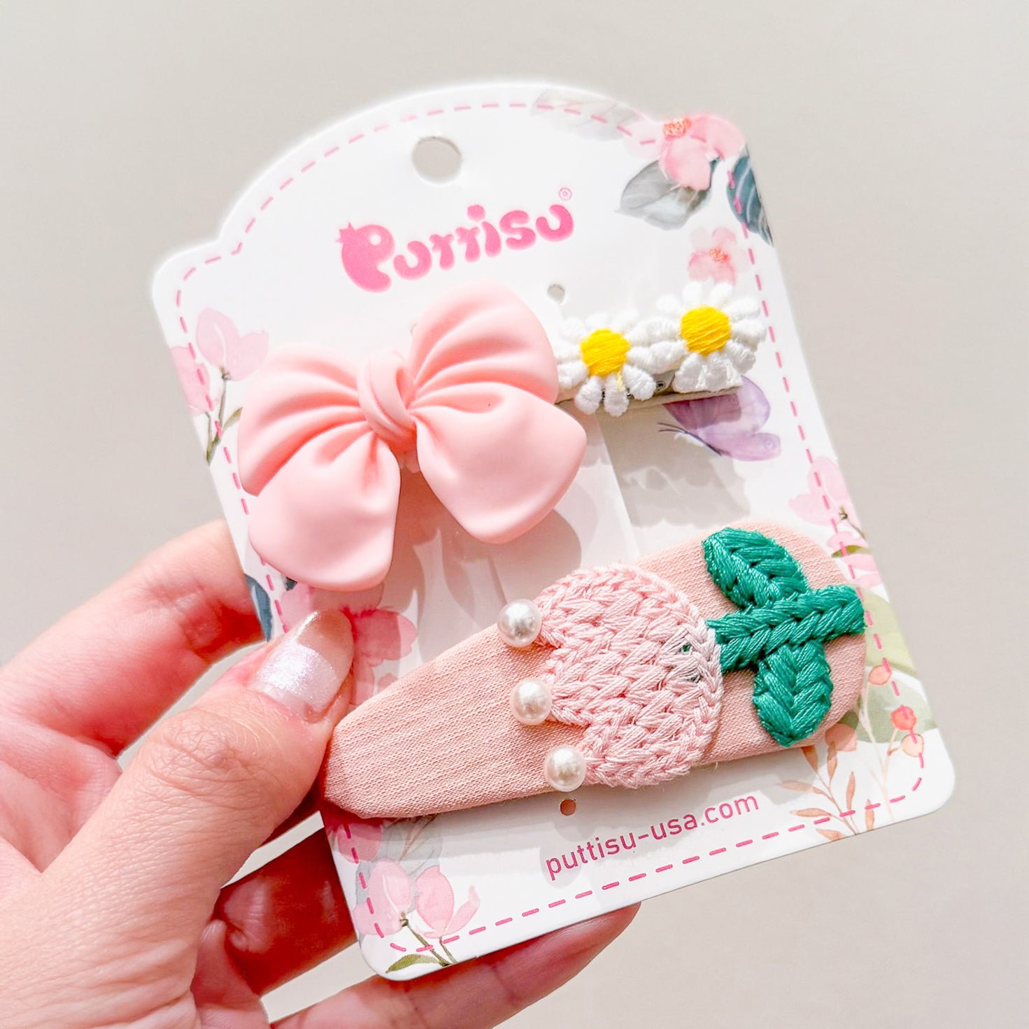 Three hair clips on a pink and white card. One with a pink bow, another with white daisies, and one with a pink embroidered design and green leaves.
