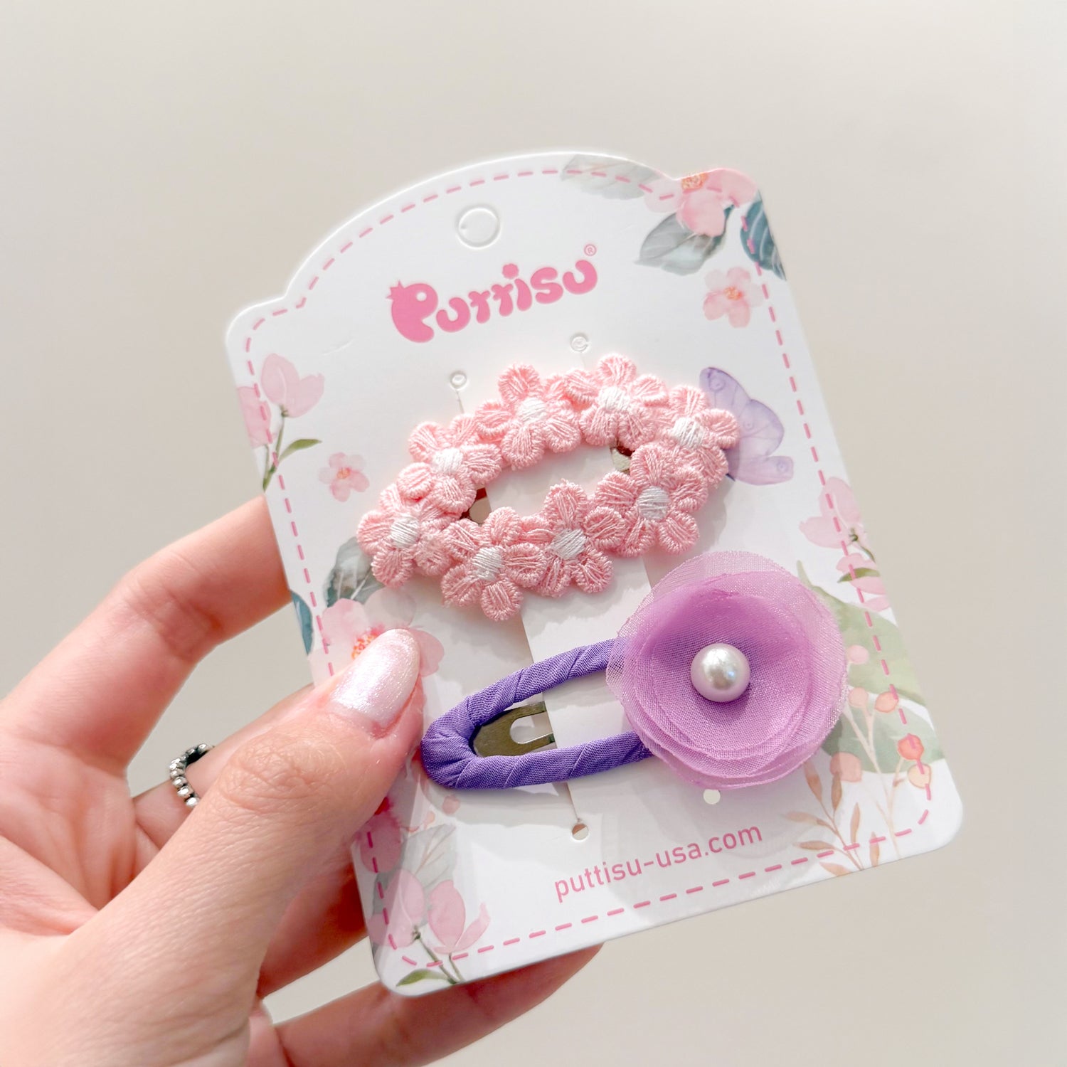 Two hair clips on a decorative card held in a hand. One pink flower hair clip and one purple flower hair clip with a pearl center.